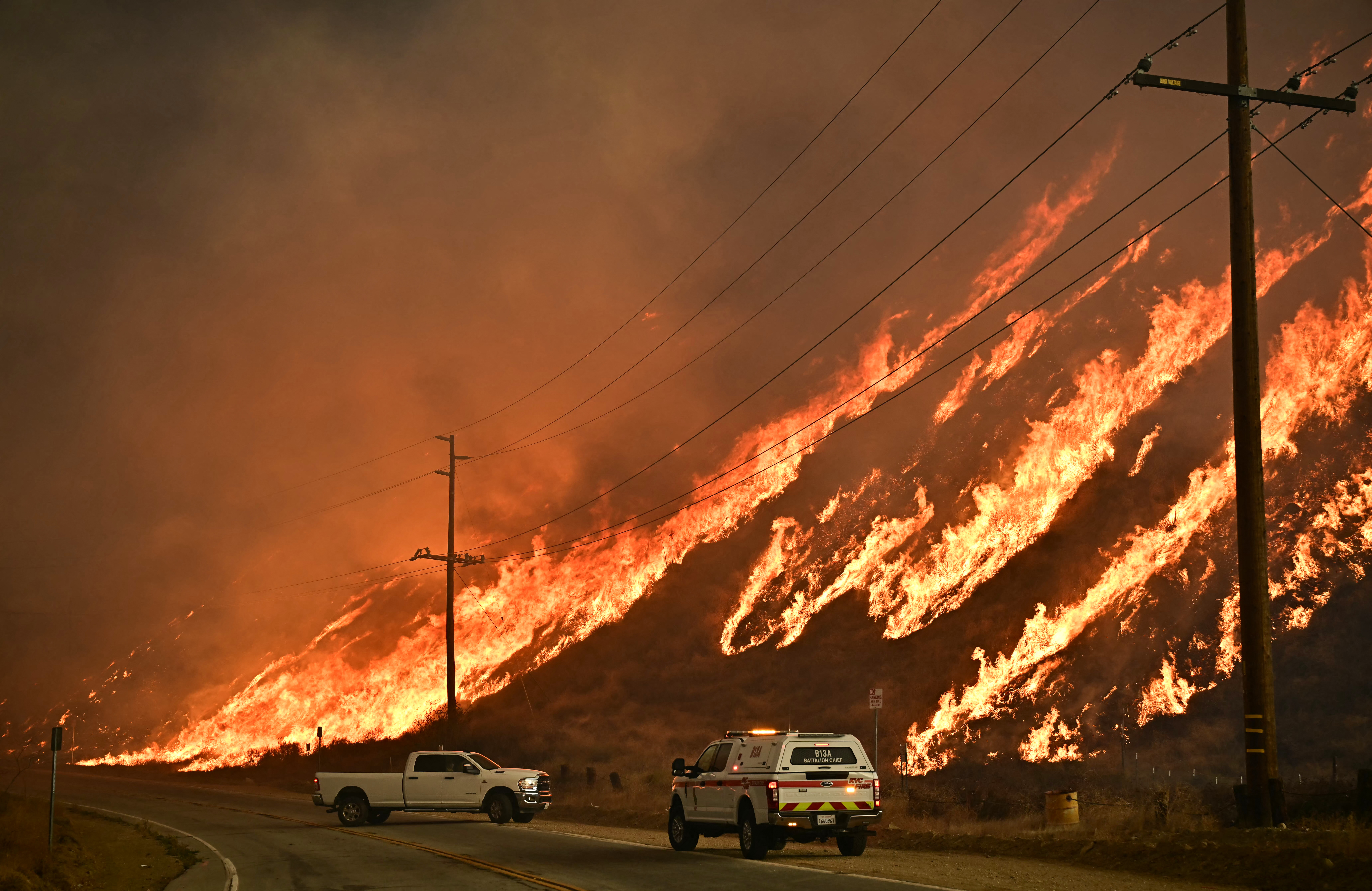 Emergency vehicles are on the side of the road as flames from the Hughes Fire race up the hill in Castaic, a northwestern neighborhood of Los Angeles, California, on January 22, 2025. A new wildfire erupted north of Los Angeles on January 22, exploding in size and sparking thousands of evacuation orders in a region already staggering from the effects of huge blazes. Ferocious flames were devouring hillsides near Castaic Lake, spreading rapidly to cover 5,000 acres (2,000 hectares) in just over two hours. The fire was being fanned by strong, dry Santa Ana winds that were racing through the area, pushing a vast pall of smoke and embers ahead of the flames. Evacuations were ordered for 19,000 people all around the lake, which sits around 35 miles north of Los Angeles, and close to the city of Santa Clarita. (Photo by Frederic J. Brown / AFP)