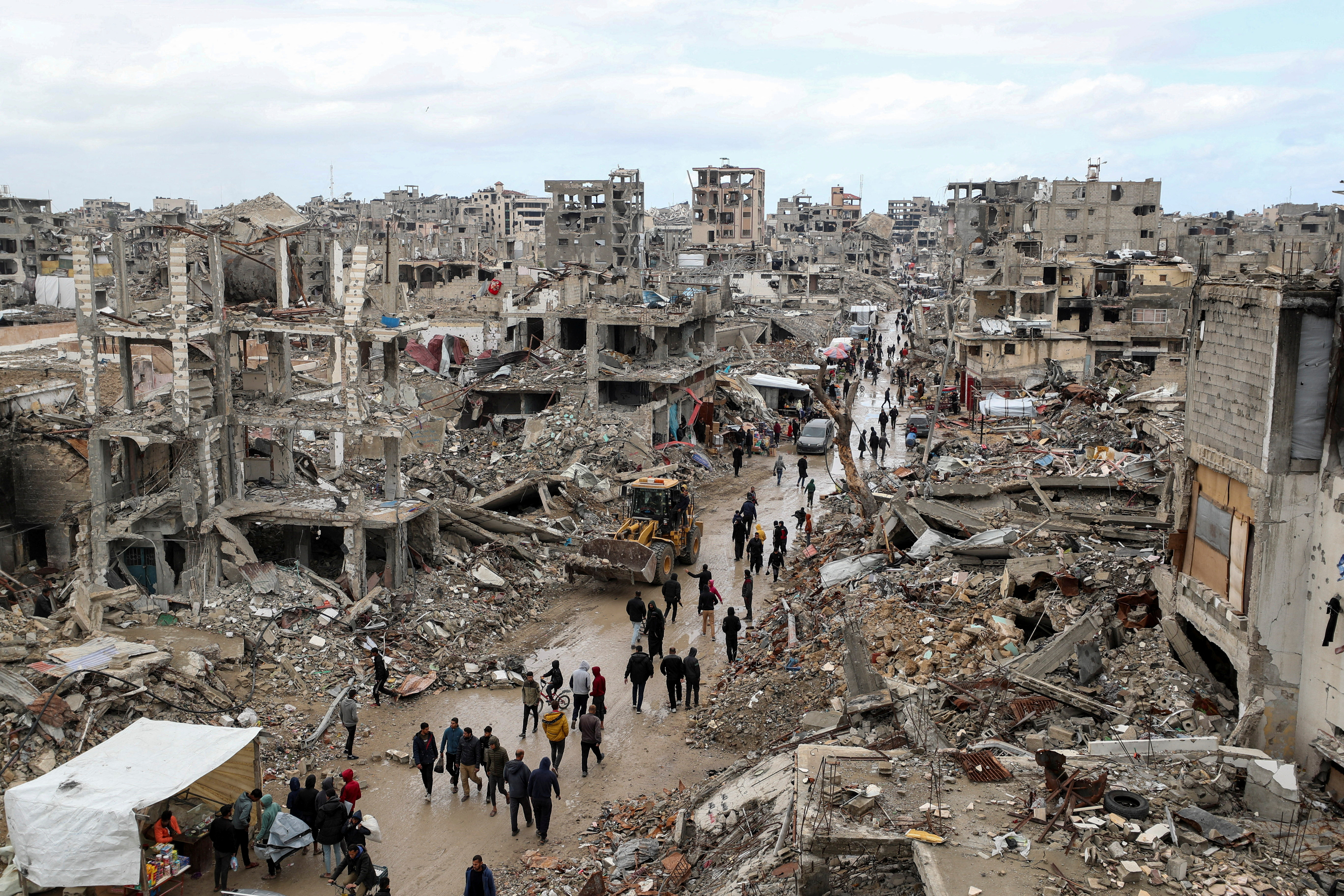 Palestinians walk past the rubble of buildings on a rainy day.