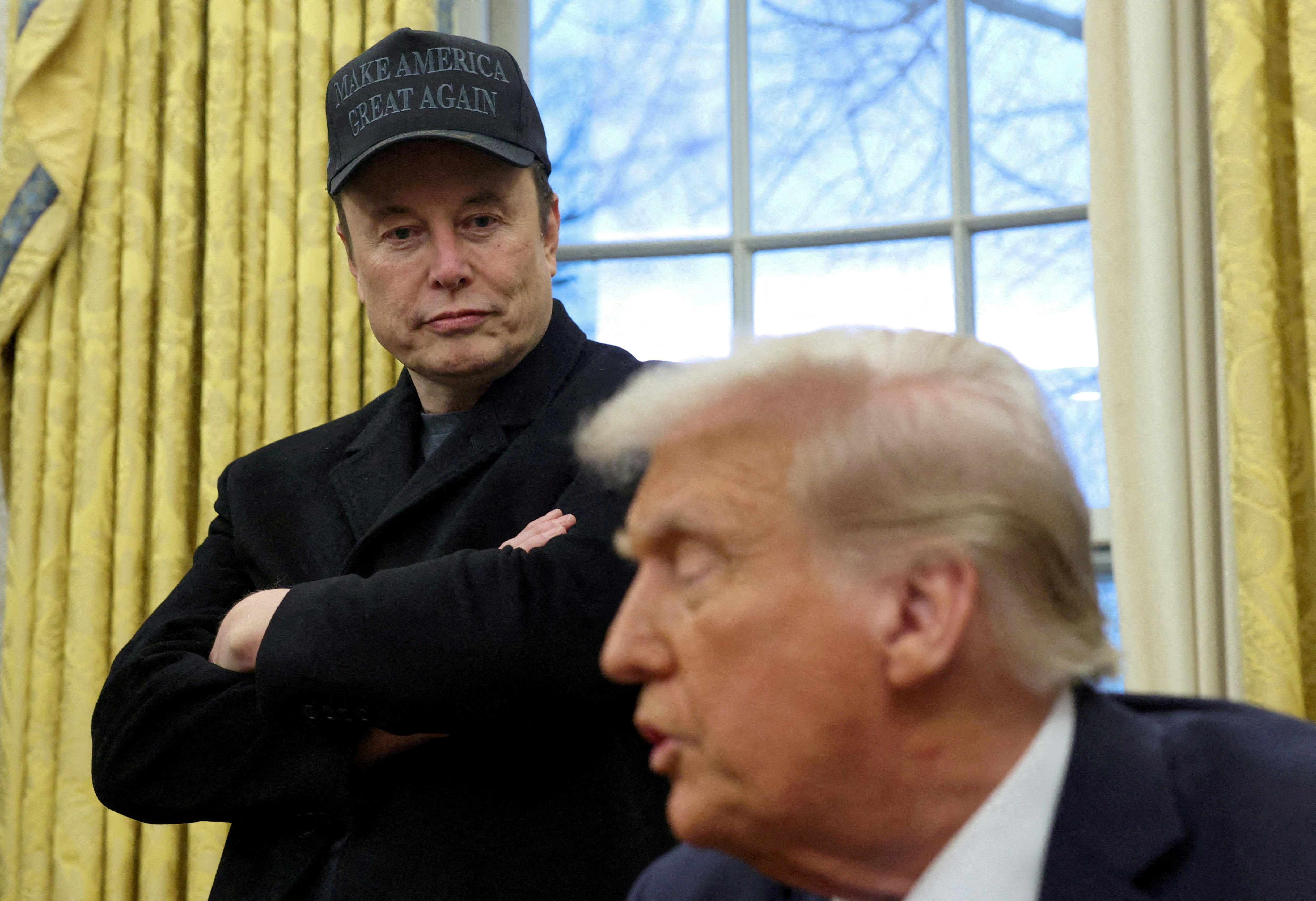 Elon Musk looks at a seated Donald Trump in the Oval Office