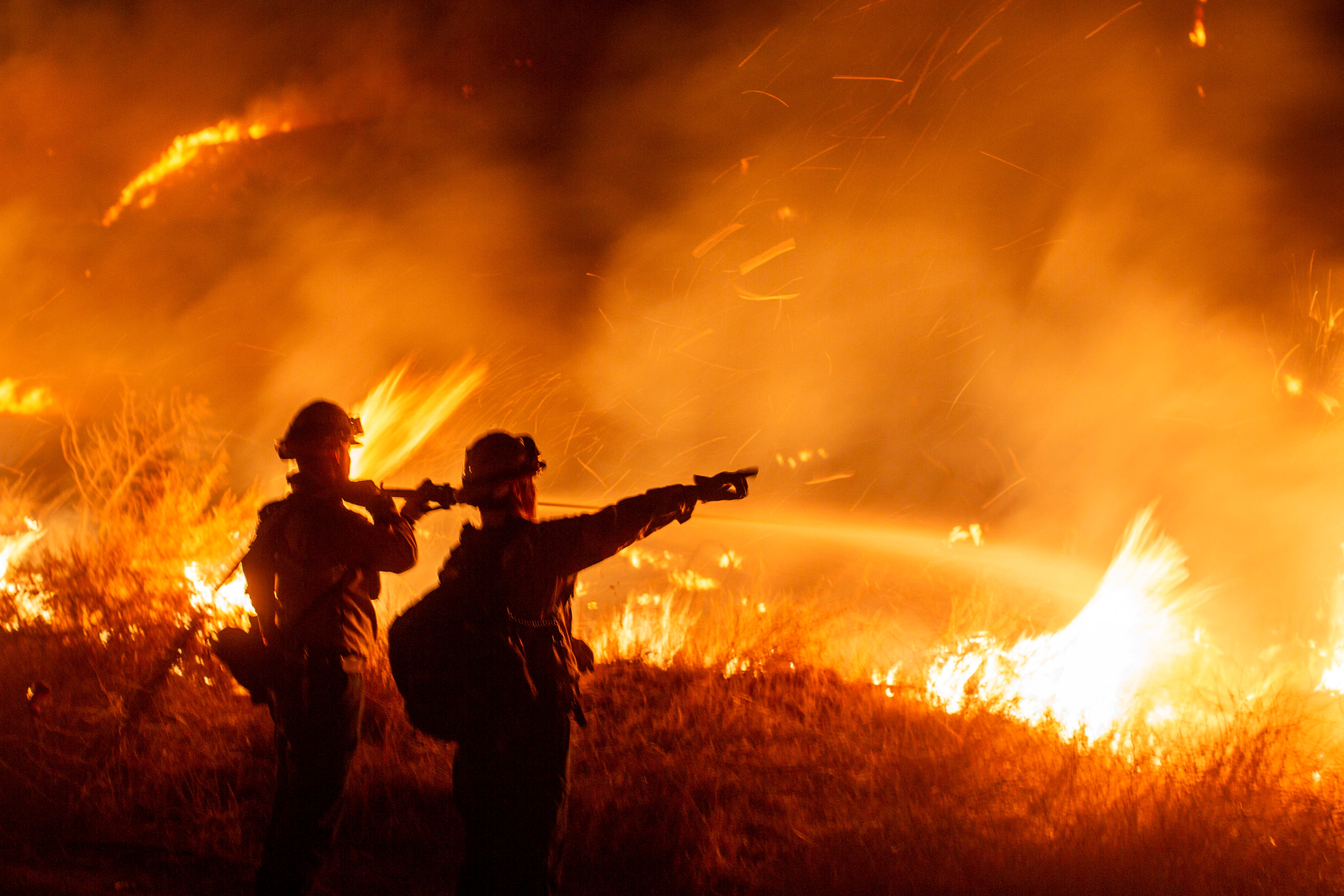 A pair of firefighters stand silhouetted against a burning blaze at night: One points.