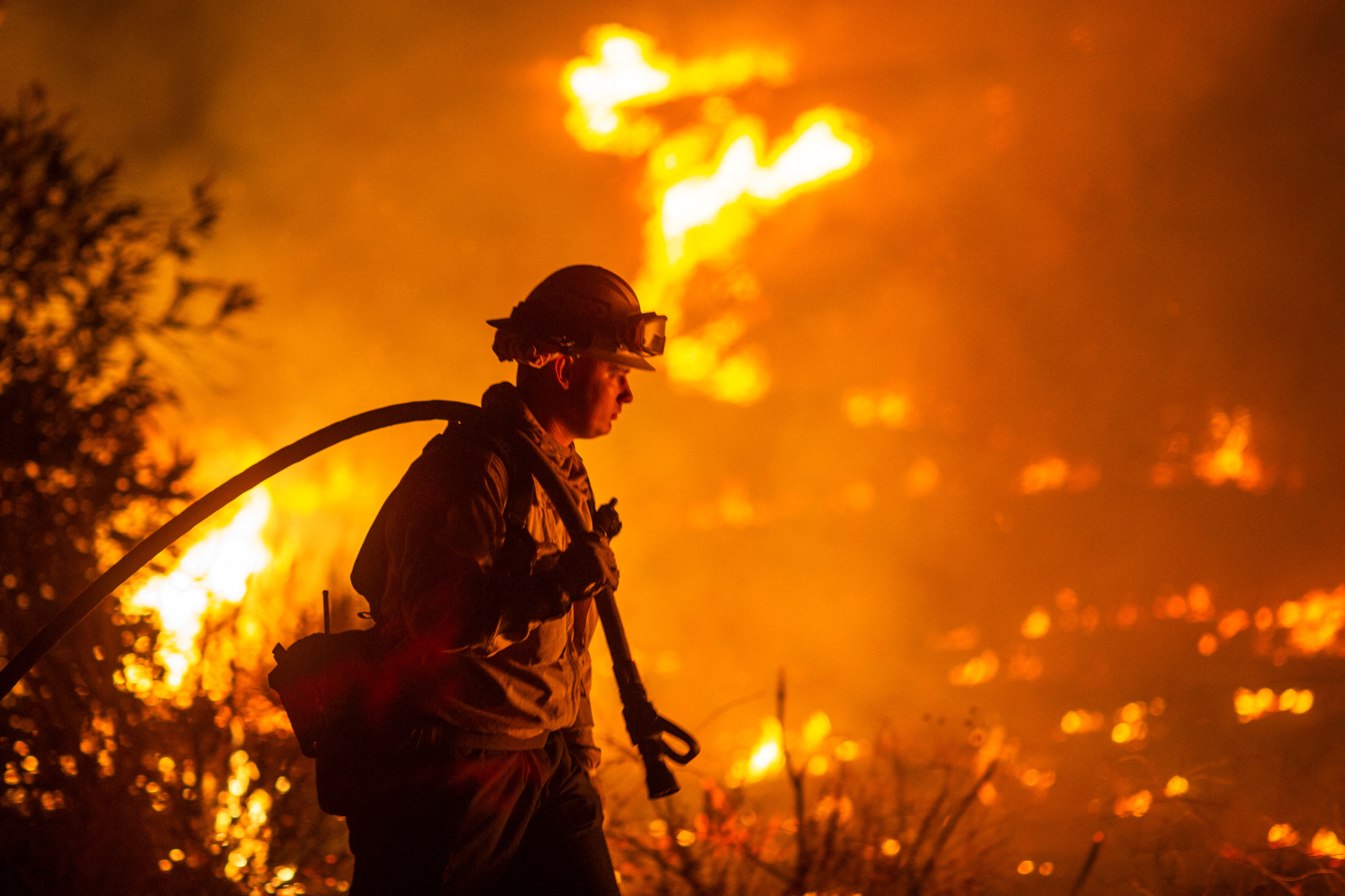 A firefighter carries a hose over their shoulder while fighting flames at night.