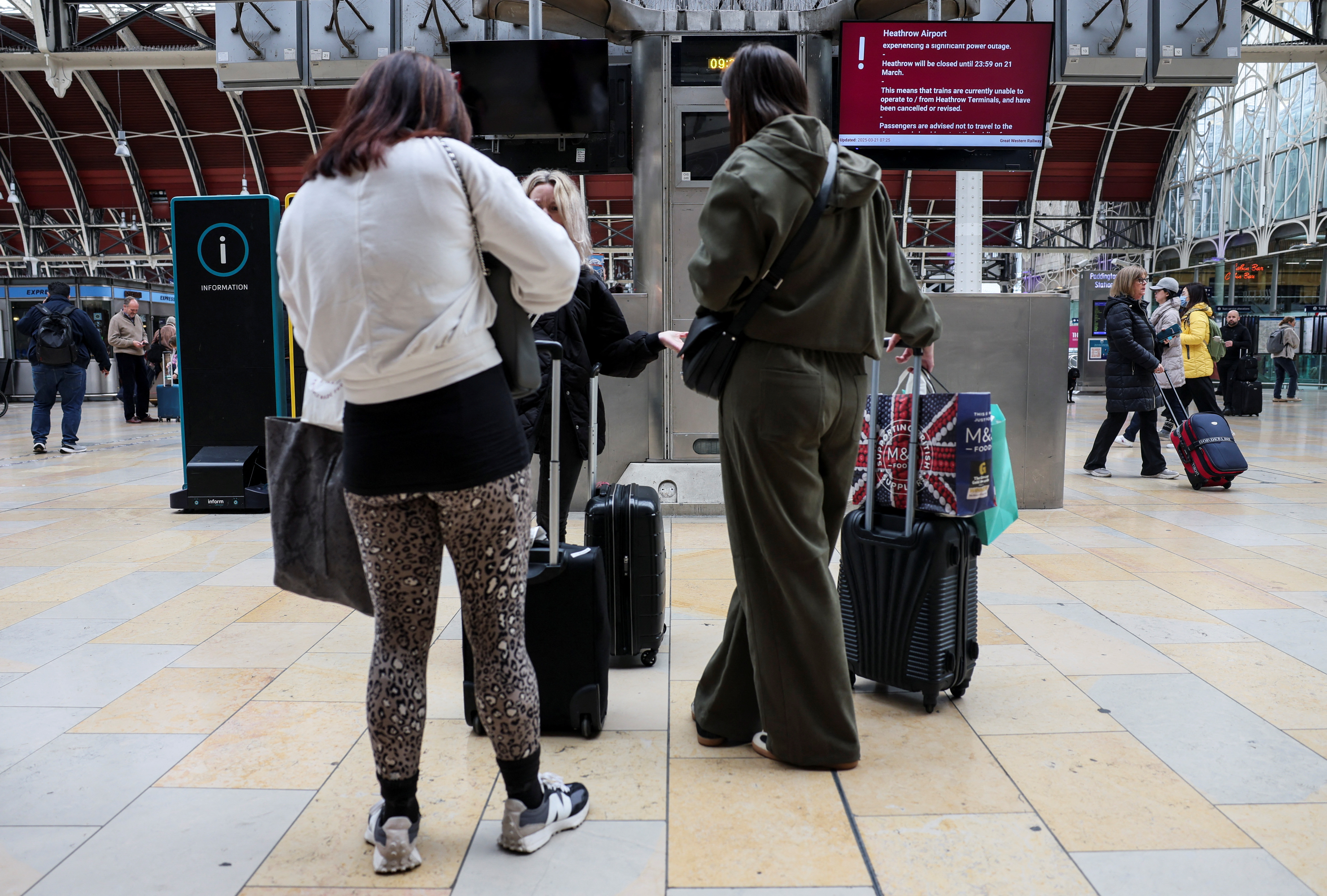 People wait at the Paddington railway station near a monitor with a notice about a power outage at Heathrow International Airport.