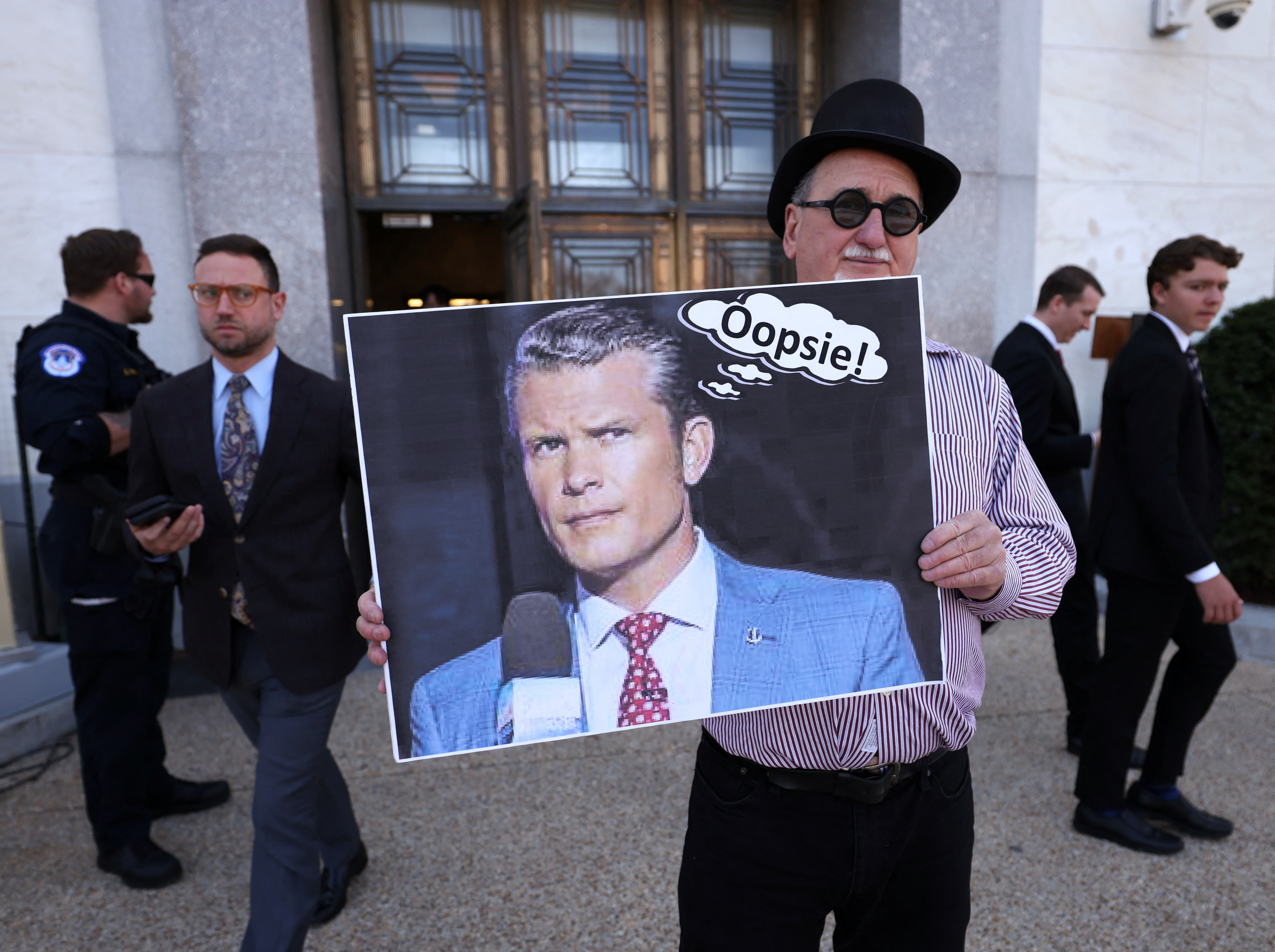 A protester holds up a picture of Pete Hegseth with a speech bubble that says, "Oopsie!"
