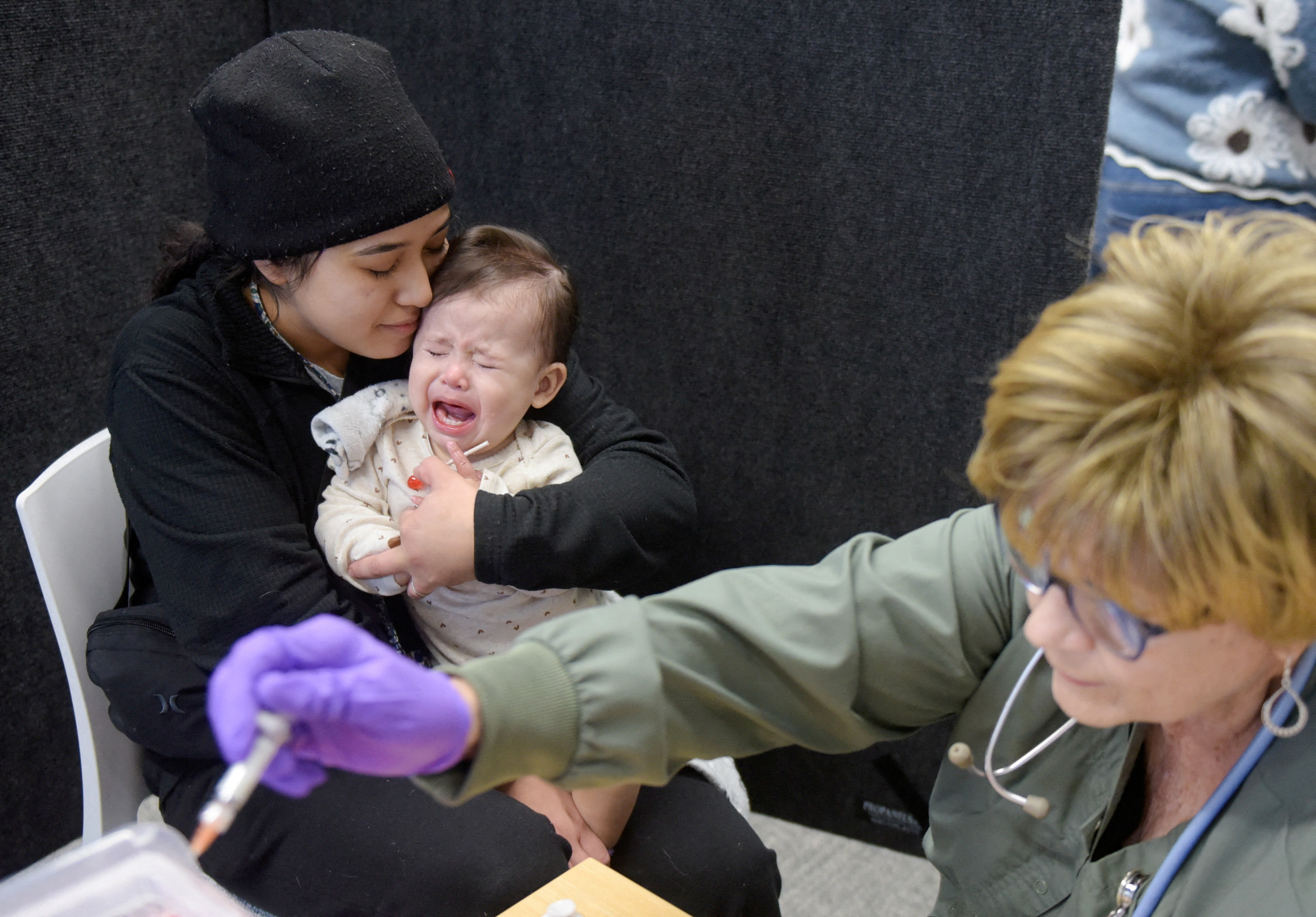 A mother holds a baby in her arms as a healthcare provider prepares a vaccine dose.