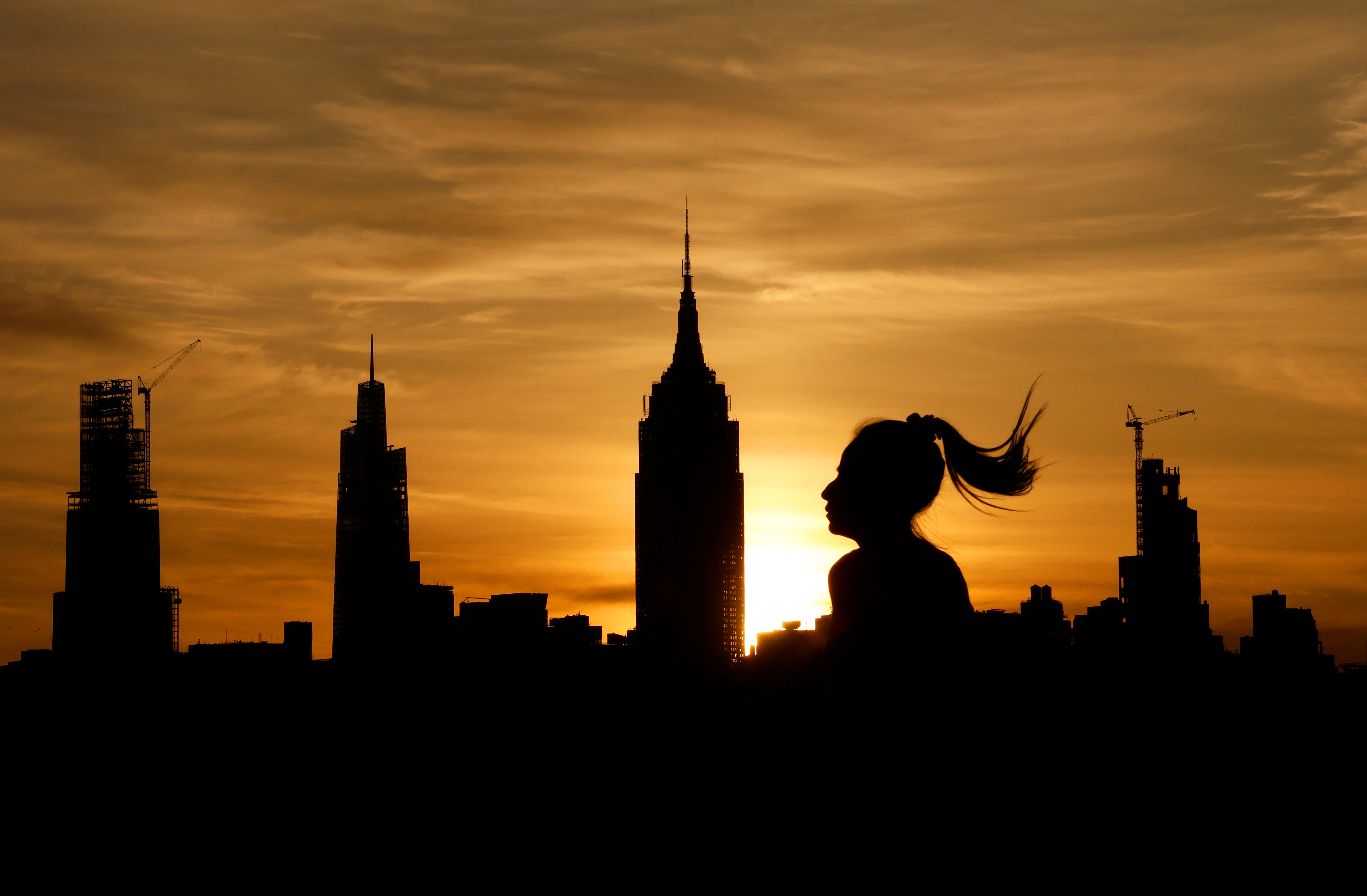 The sun rises behind the skyline of midtown Manhattan and the Empire State Building in New York City as a woman runs in a park.