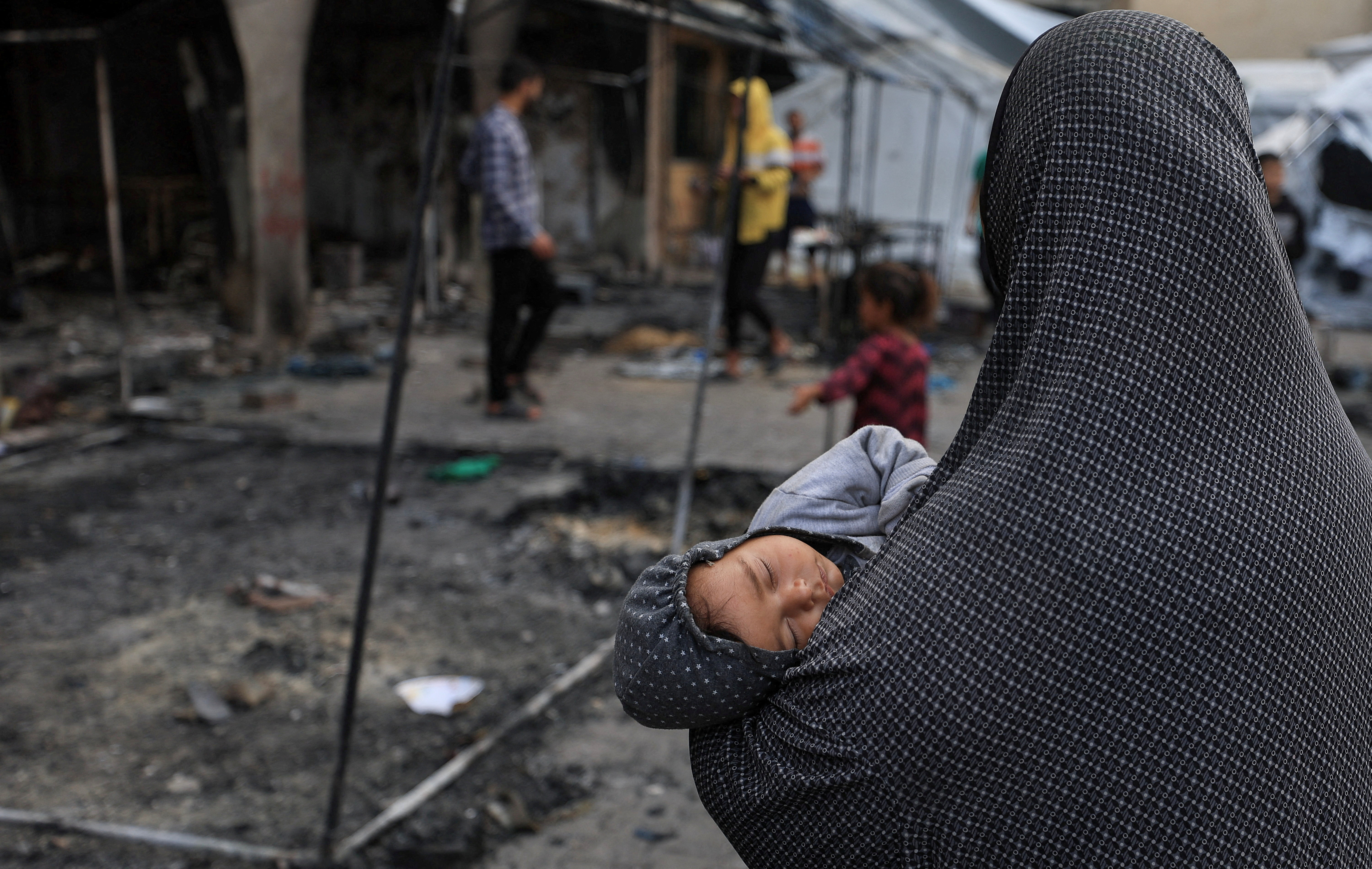 A Palestinian woman holds a child while inspecting the damage at a school sheltering displaced people, following an Israeli attack.