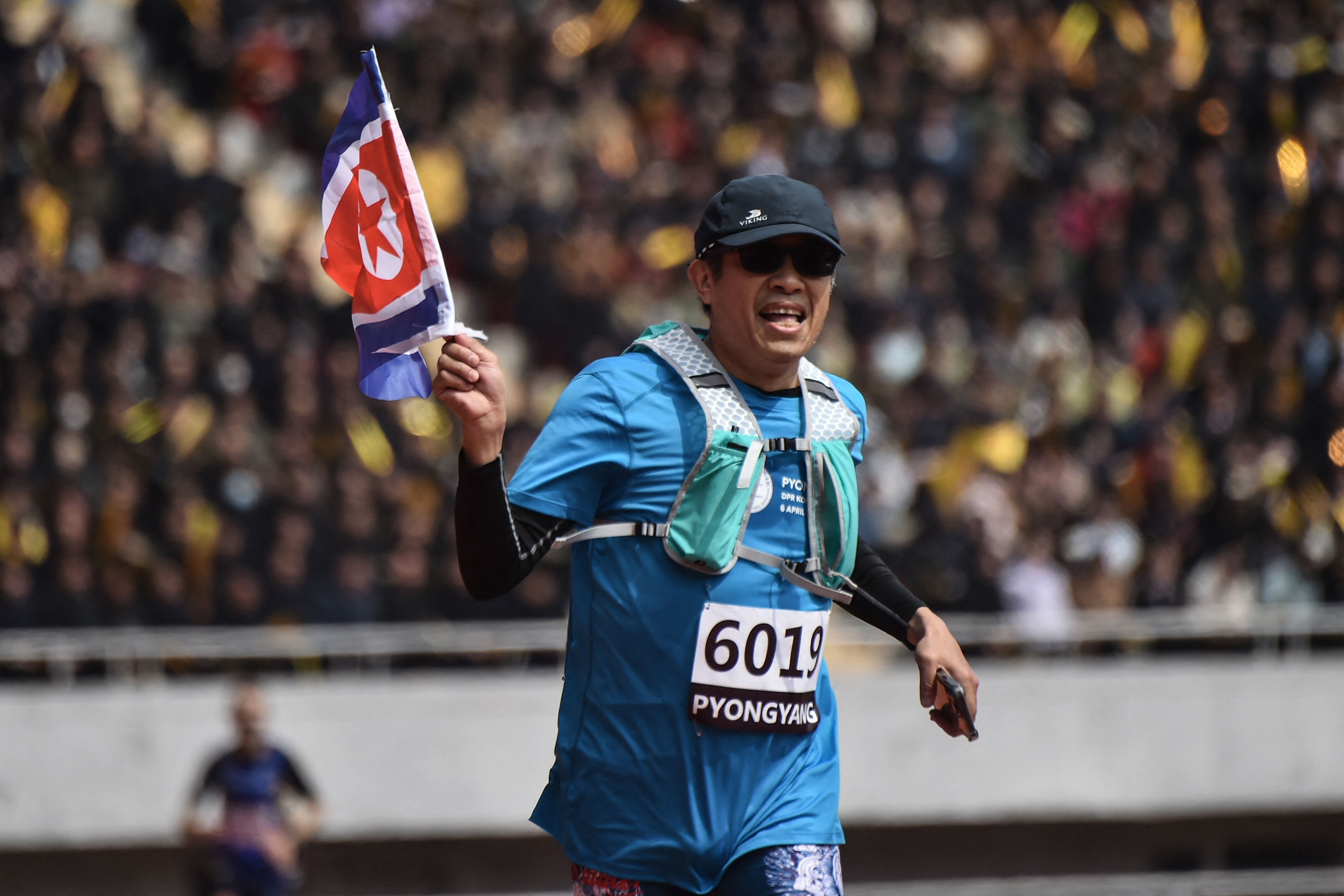 A man runs holding a North Korean flag. He is in focus and the crowd in the stands is blurred behind him.