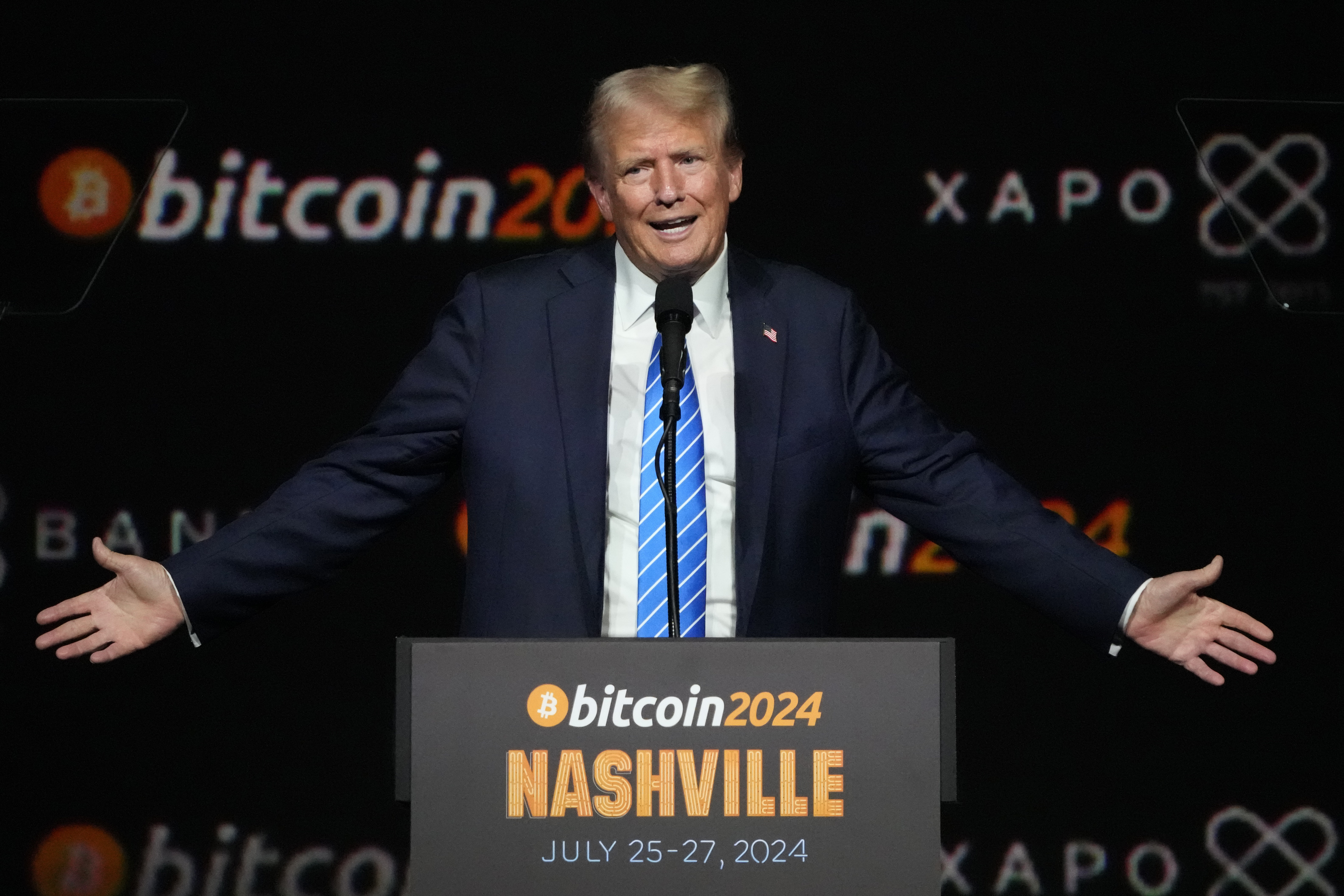 Donald Trump gestures, arms outstretched, behind a Bitcoin Nashville podium.