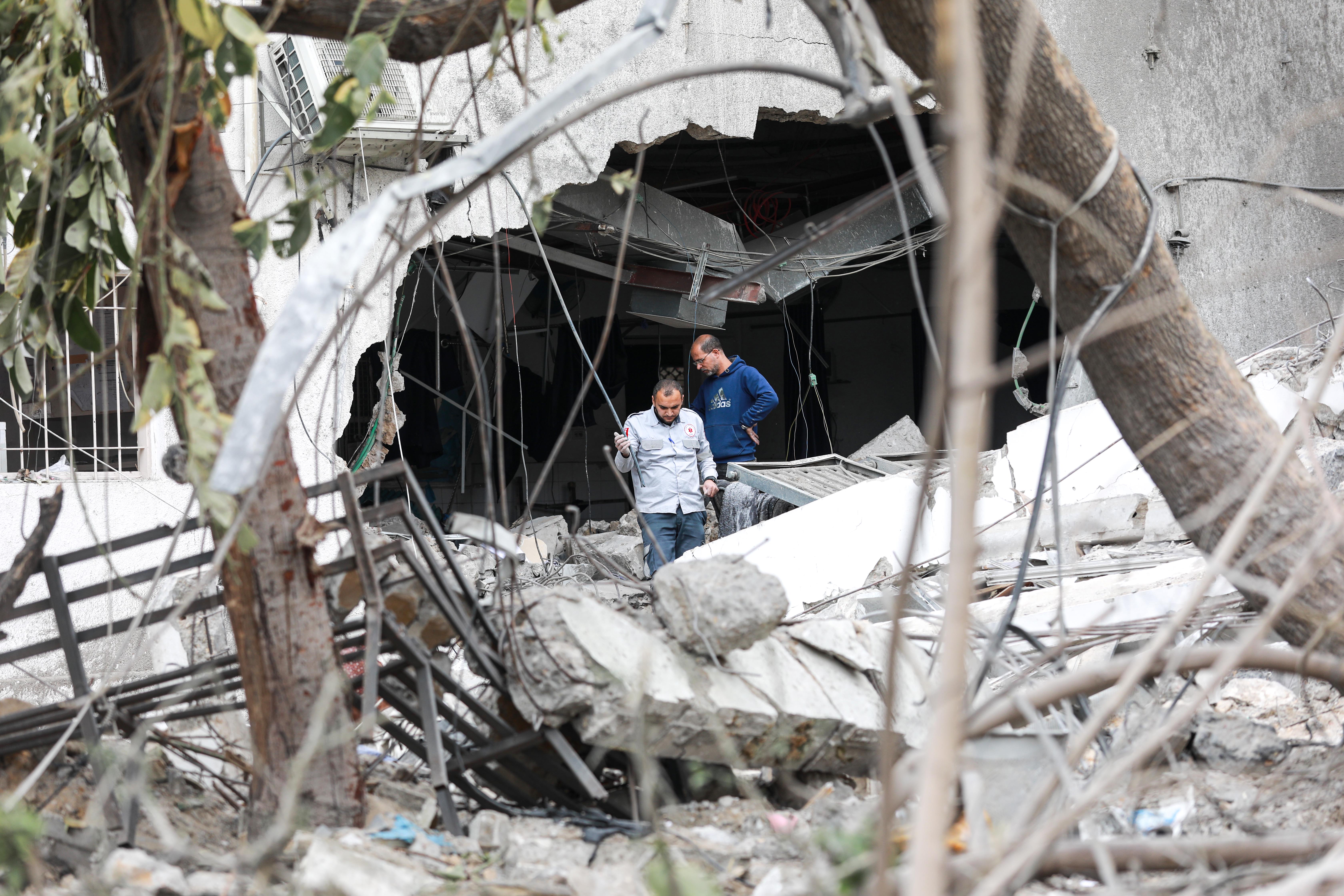 Two men walking through debris out of a huge gaping hole in the side of a building, HVAS tubes are dangling from the ceiling