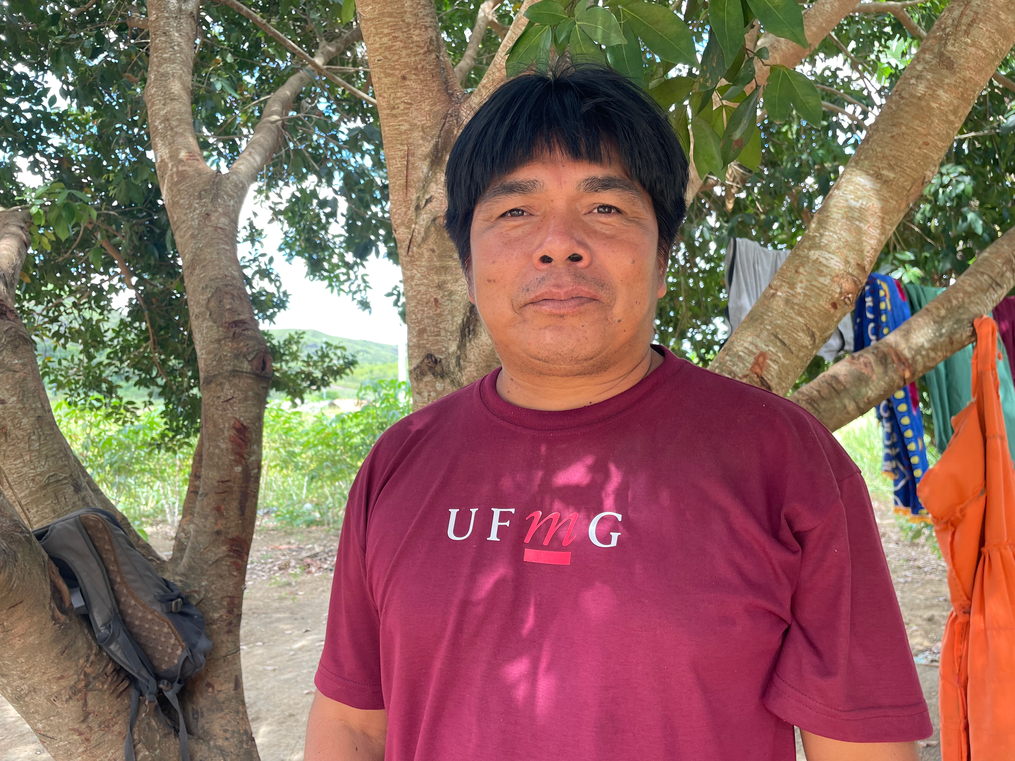 A man in a burgundy shirt stands under a tree outdoors in Brazil.