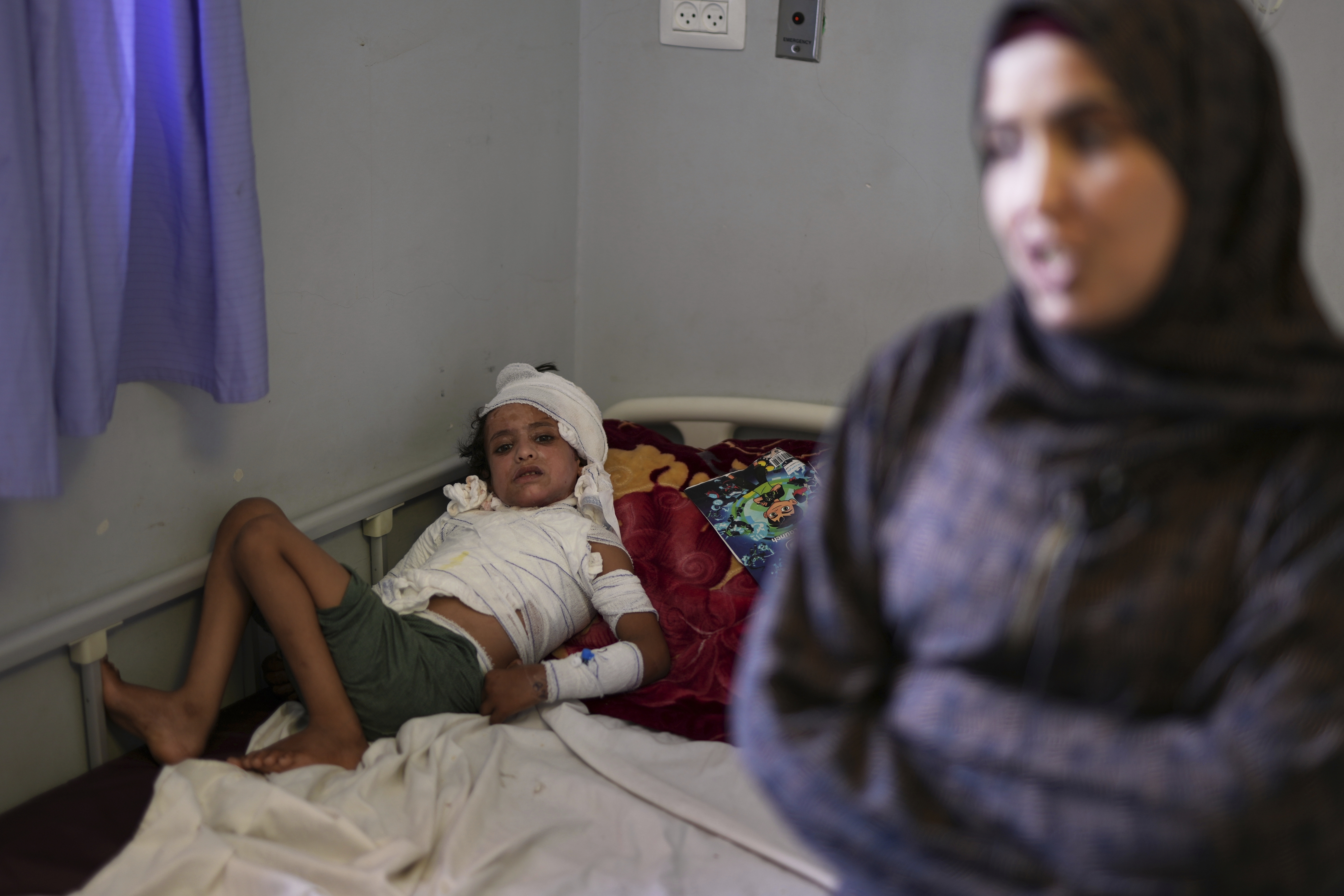 A boy who is nearly completely wrapped in bandages, except for on his legs and face, is lying on a hospital bed. He is pressed against the wall and looks wincing at the camera.