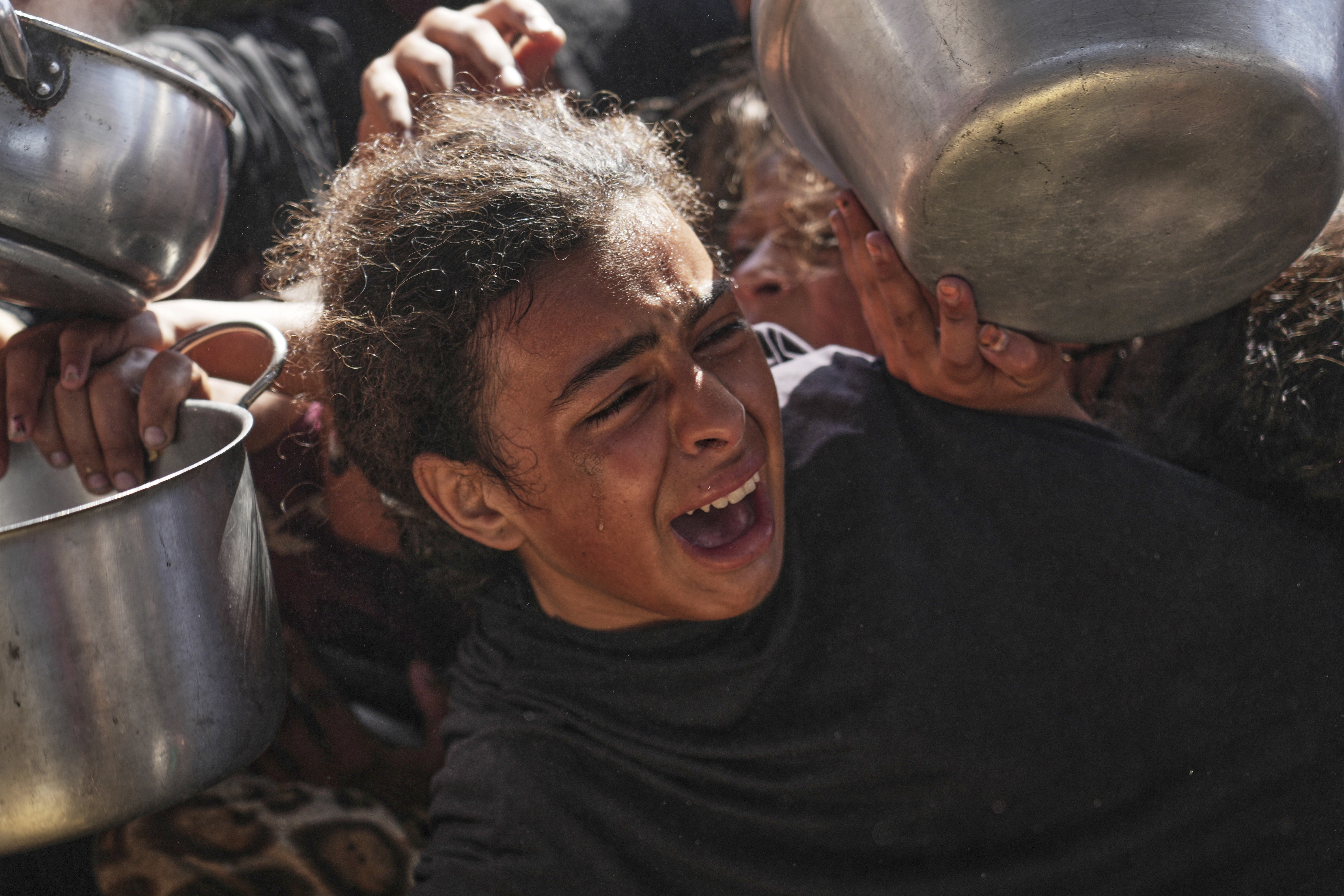 A girl is crying with her mouth open while there are pans and pots all around her, held up by the arms of others wanting food.
