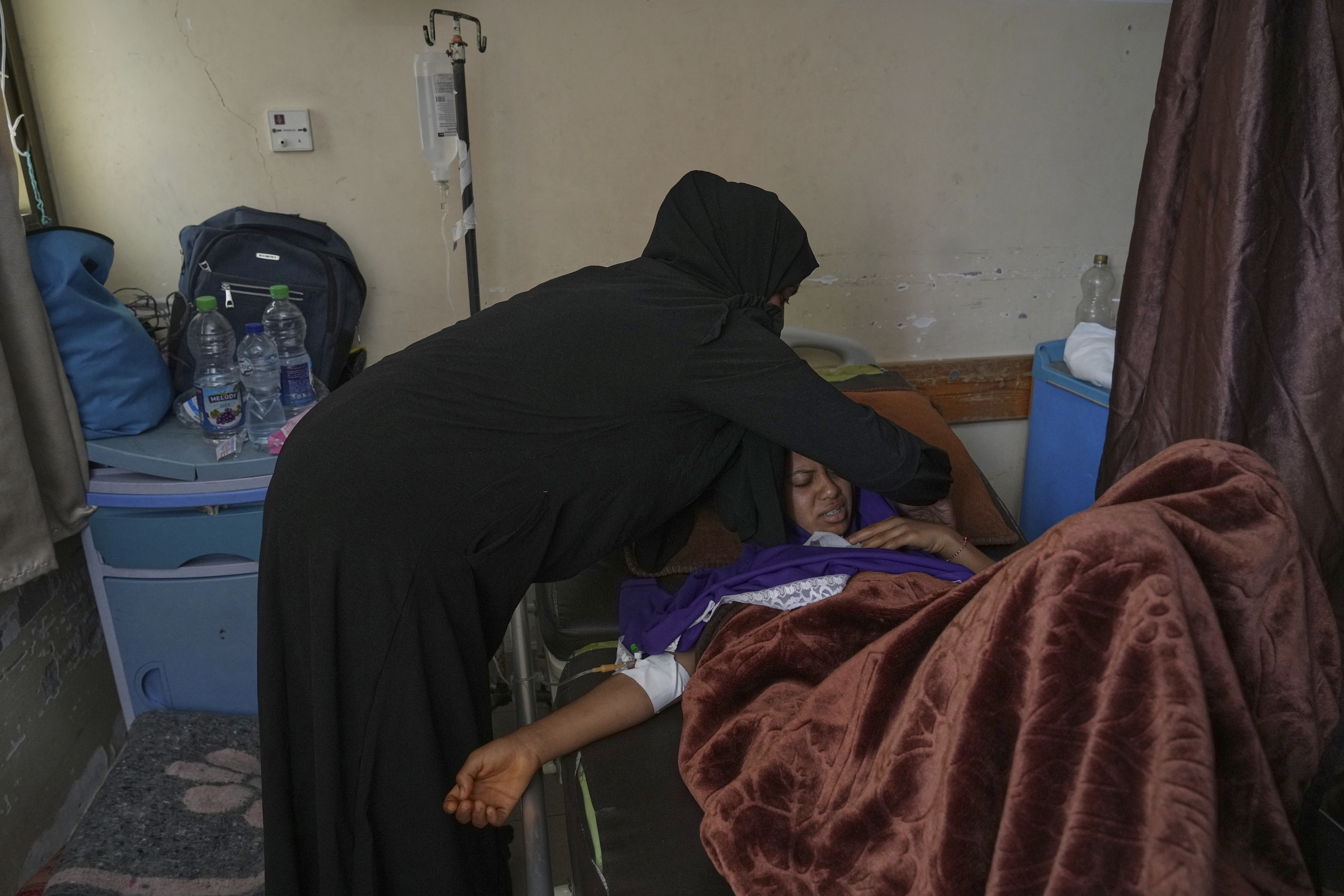 A woman leans over a young woman lying in bed and wincing in pain. She has a brown blanket on her.