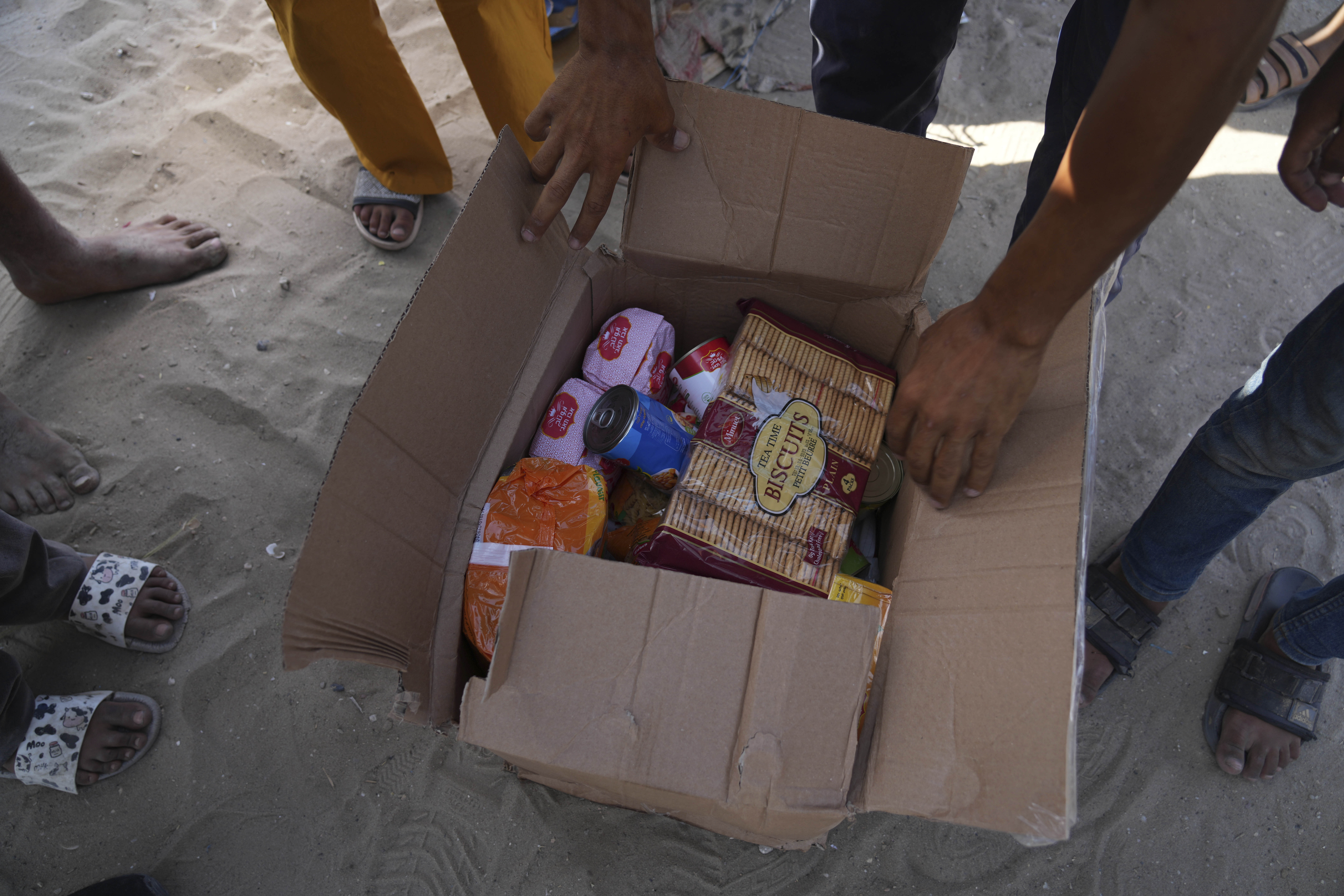 Palestinians reach into an open cardboard box of aid, featuring "Teatime biscuits" and cans of food.