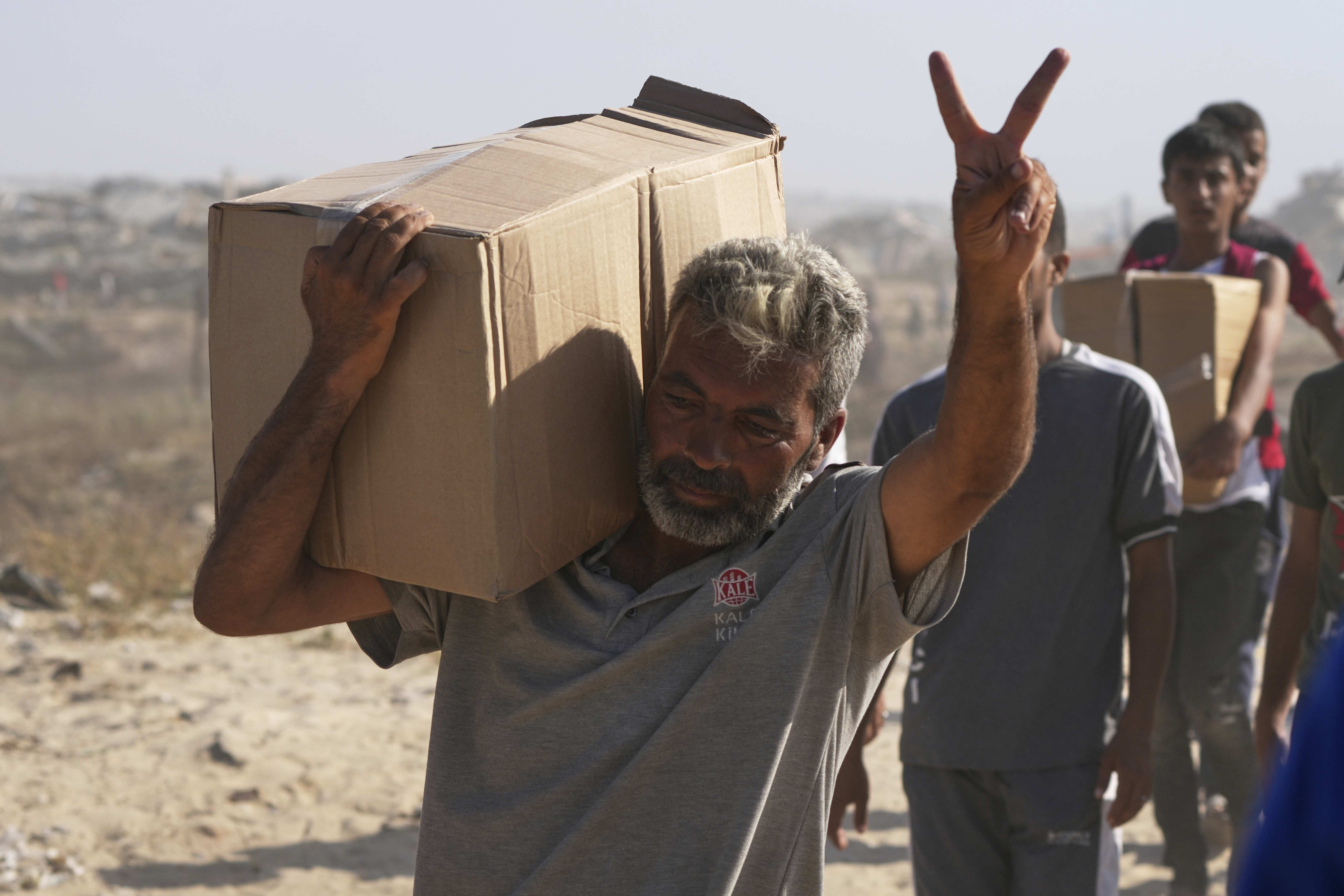 A bearded man carries a cardboard box over his shoulder, flashing a peace sign with his free hand.