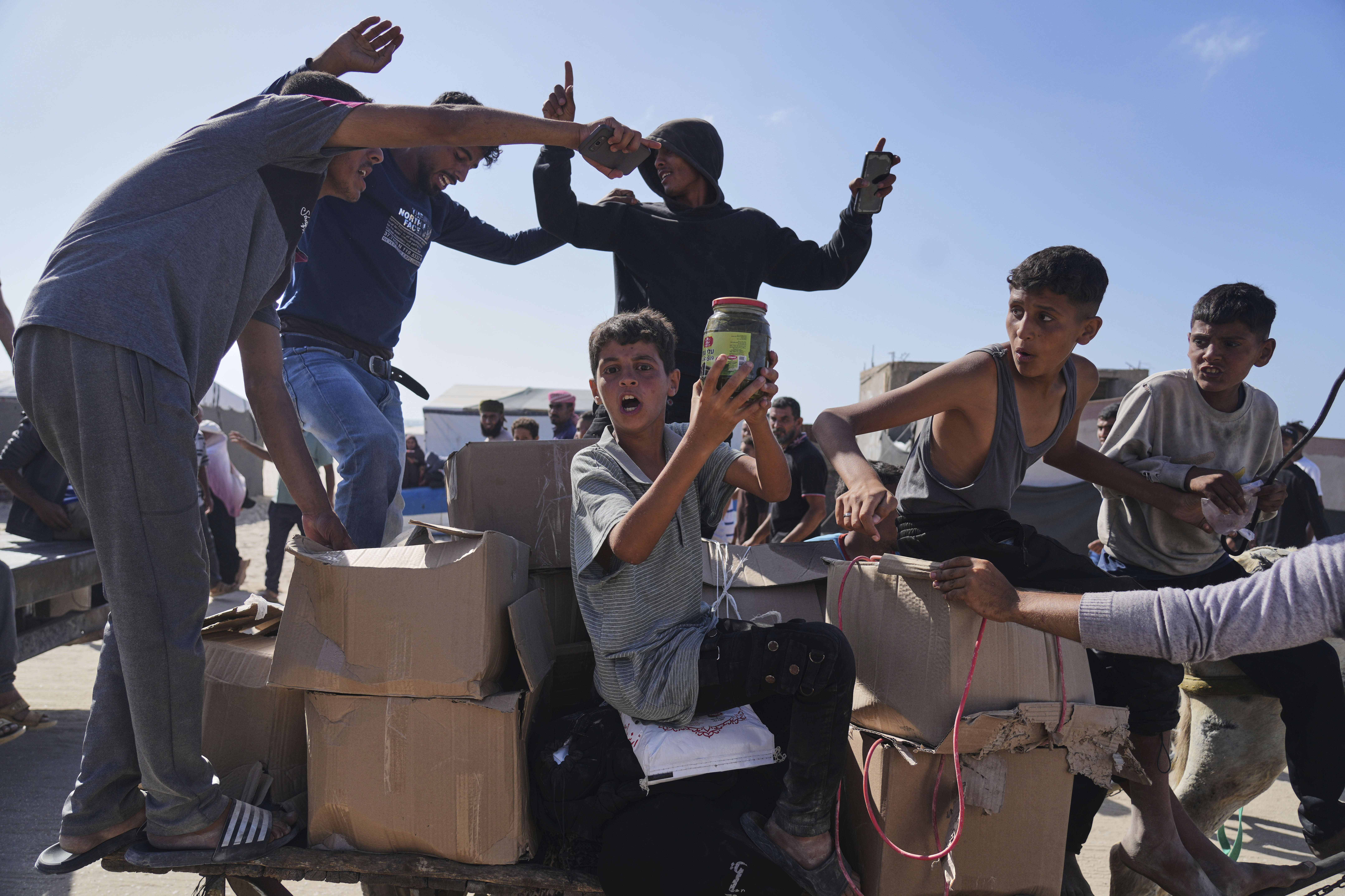 A boy holds up a glass jar of food as he sits on the back of a cart filled with cardboard boxes. The men and boys around him gesture, some with their arms in the air.