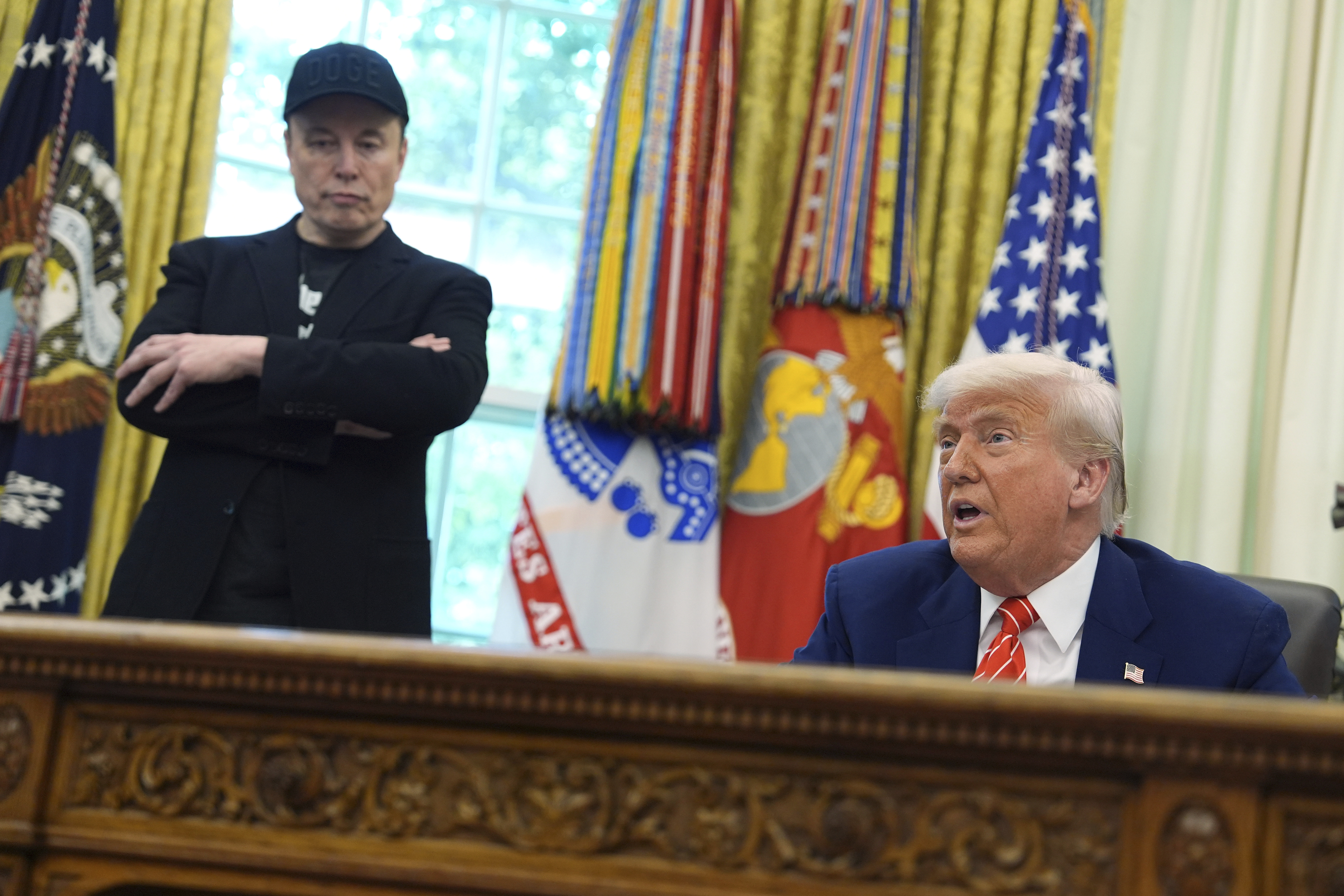 Elon Musk, dressed in black, stands with his arms crossed behind the Resolute Desk while Donald Trump, seated, speaks.