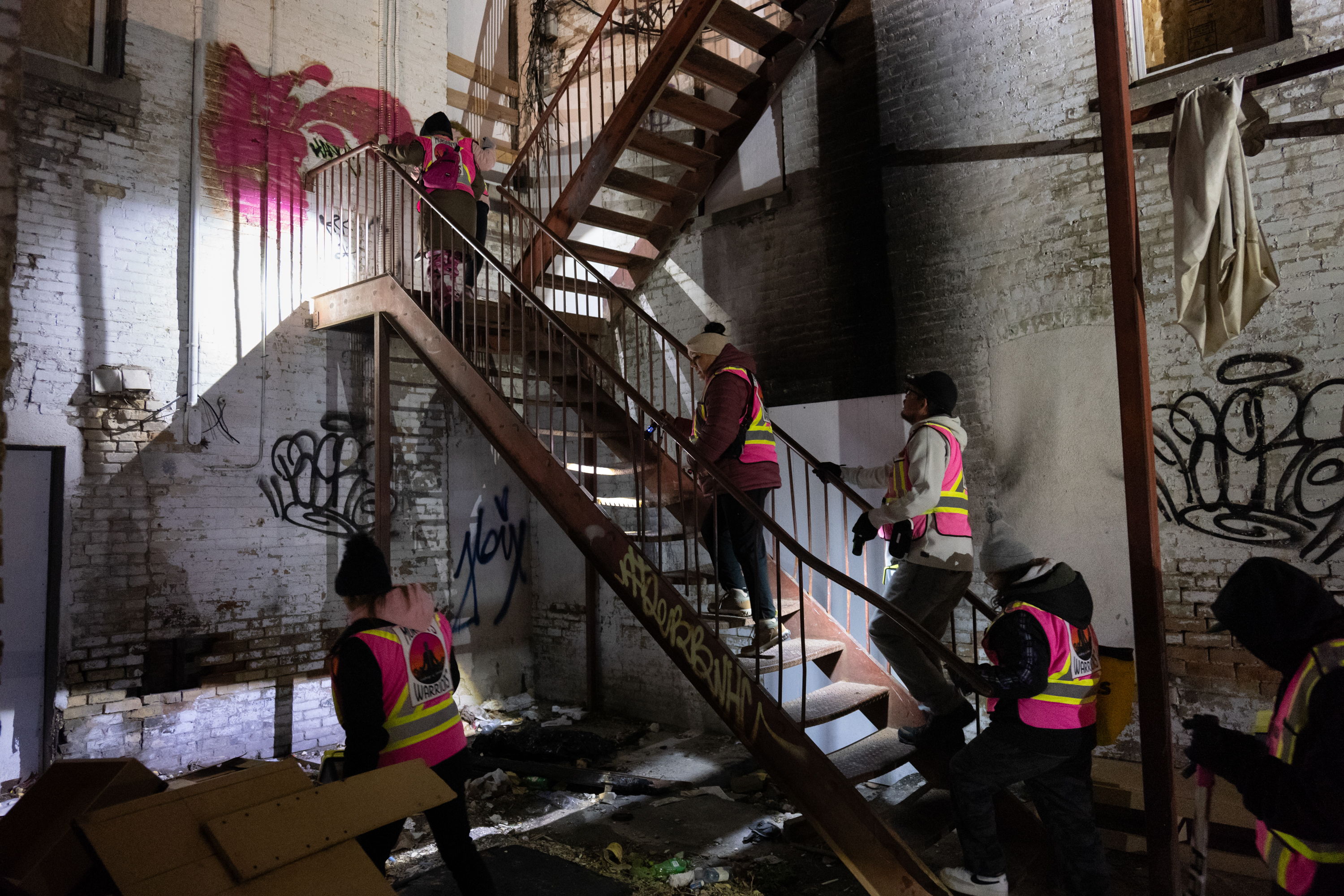 People in fluorescent pink vests climb a metal staircase inside a white brick building with graffiti on the walls