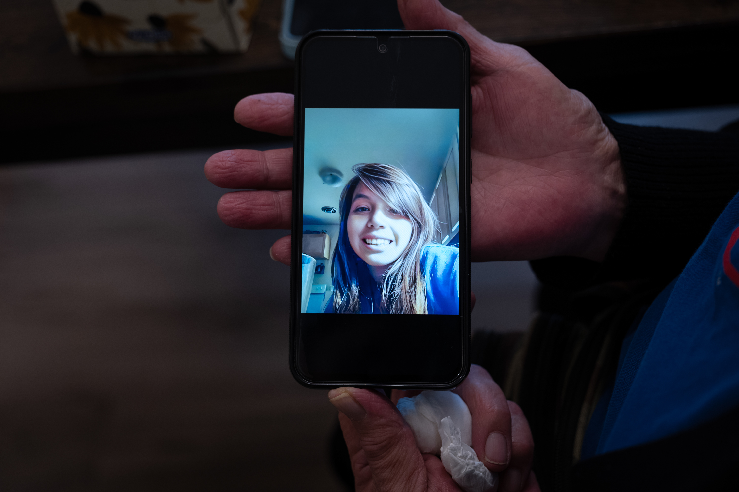 A hand holds a mobile phone. On the phone is a photo of a young woman with long dark hair