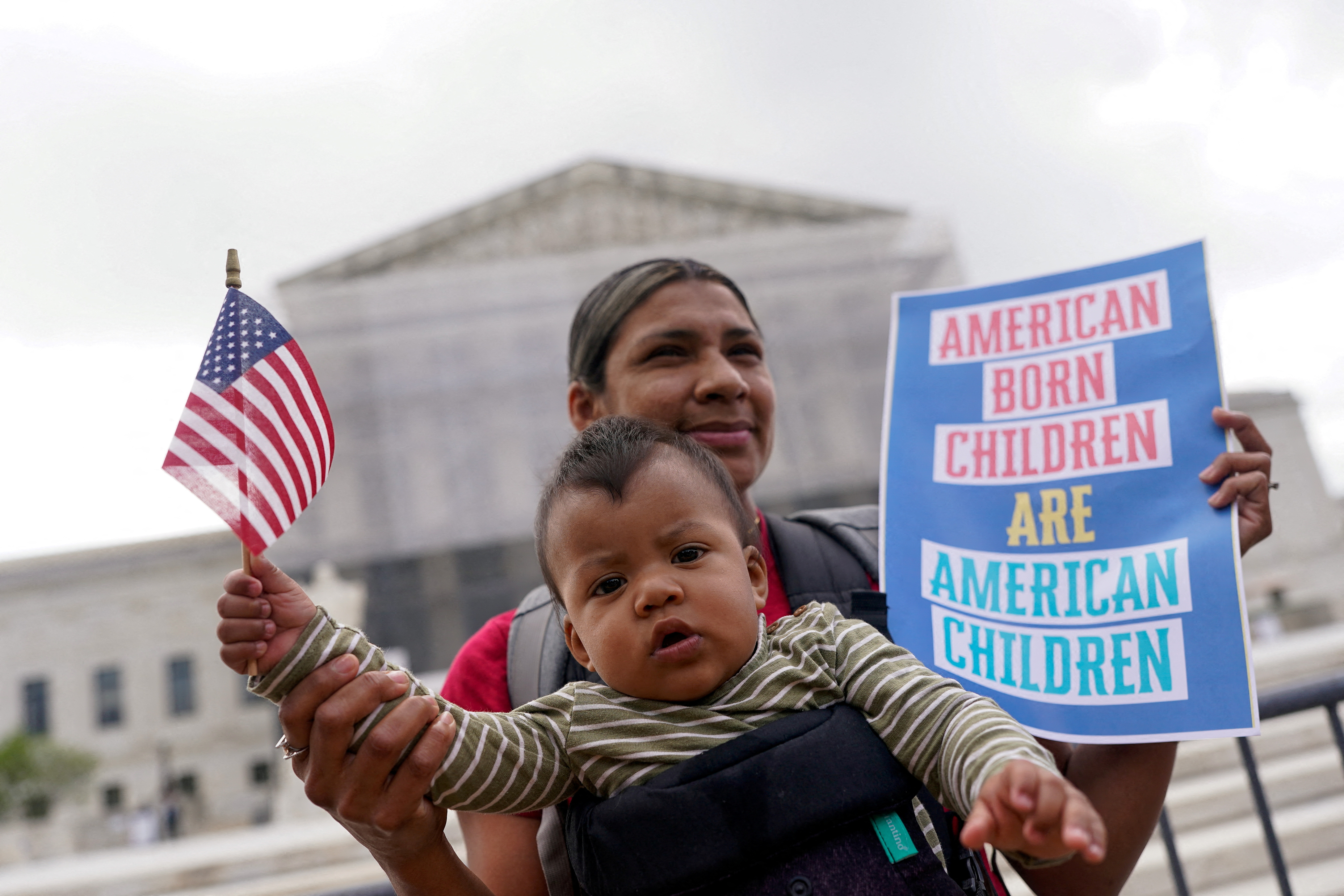 A woman with a baby strapped to her chest holds a sign as she stands before the Supreme Court: "American born children are American children."