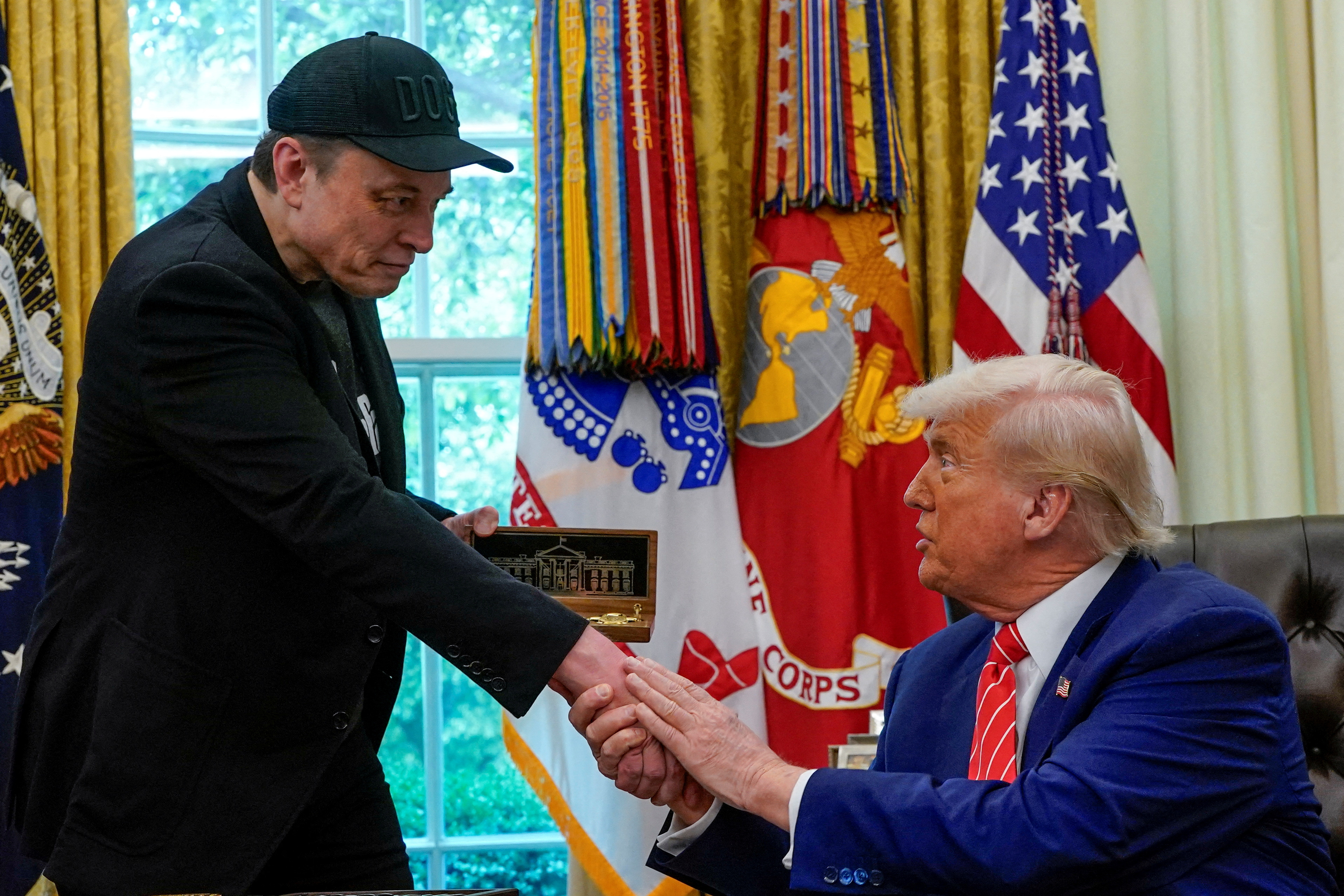 Elon Musk leans over to shake hands with a seated Donald Trump in the Oval Office.