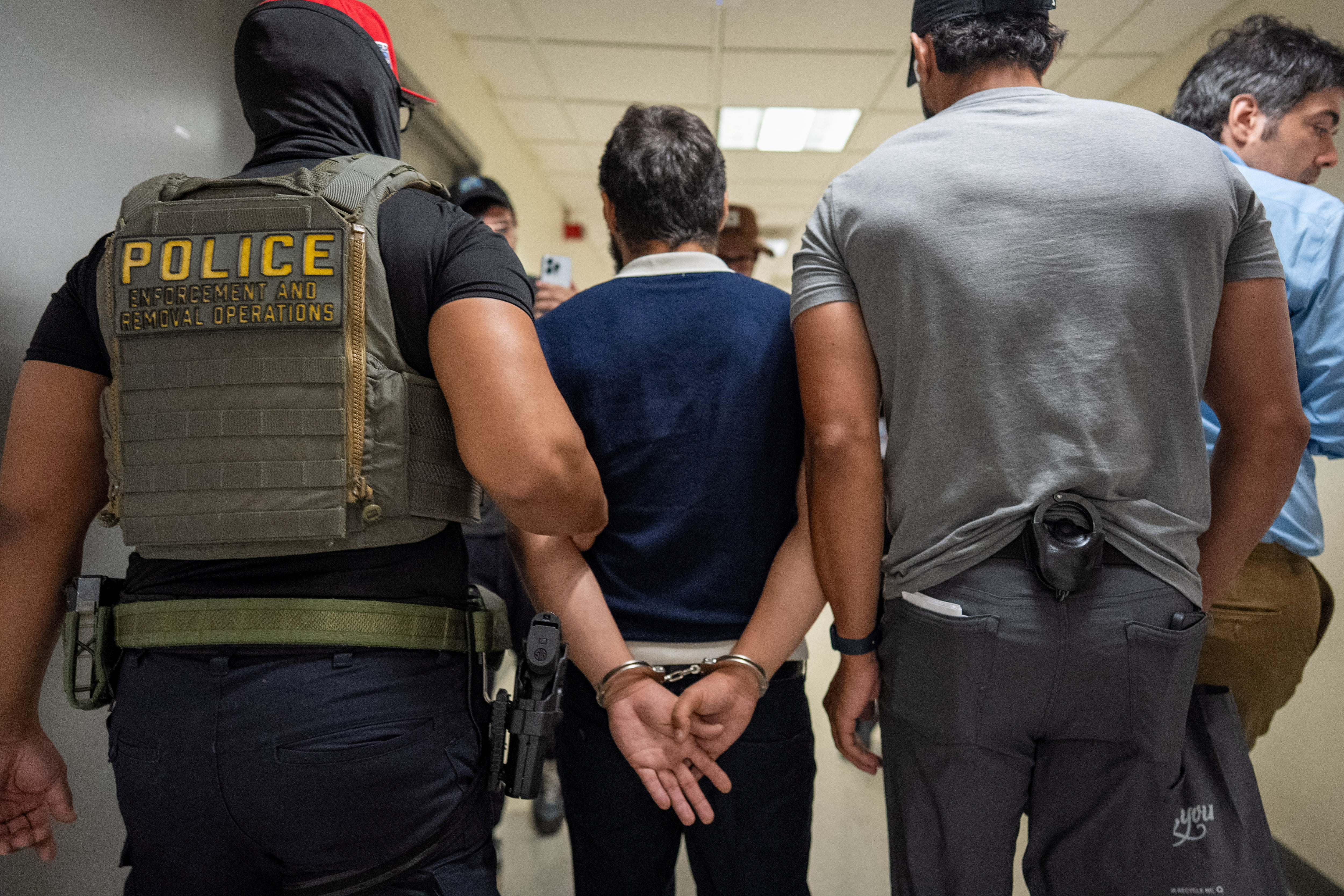 Federal immigration agents stand on either side of a migrant man they are leading away in handcuffs.