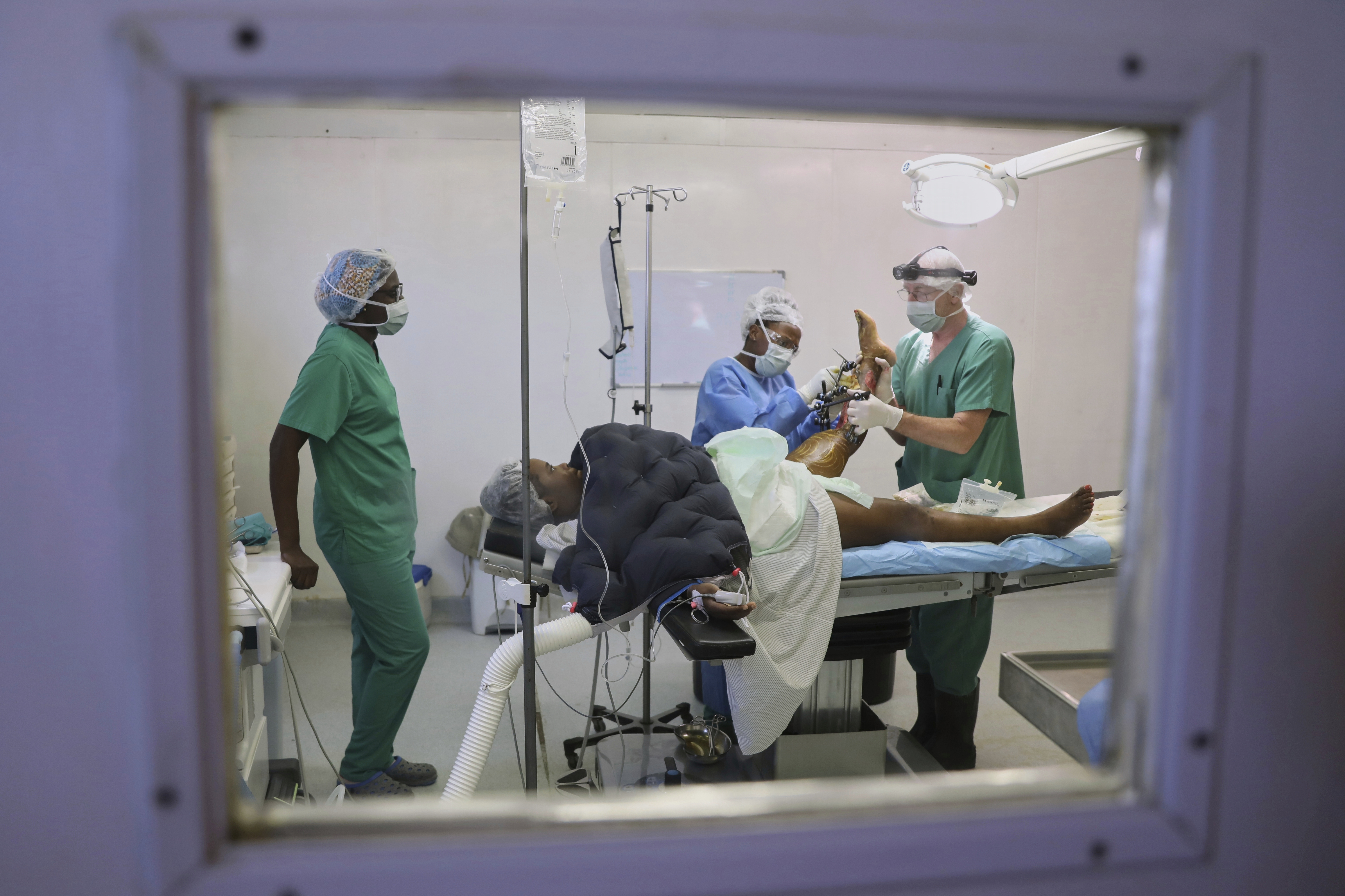 Healthcare workers — seen through a window — lift the foot of a patient on a hospital bed.