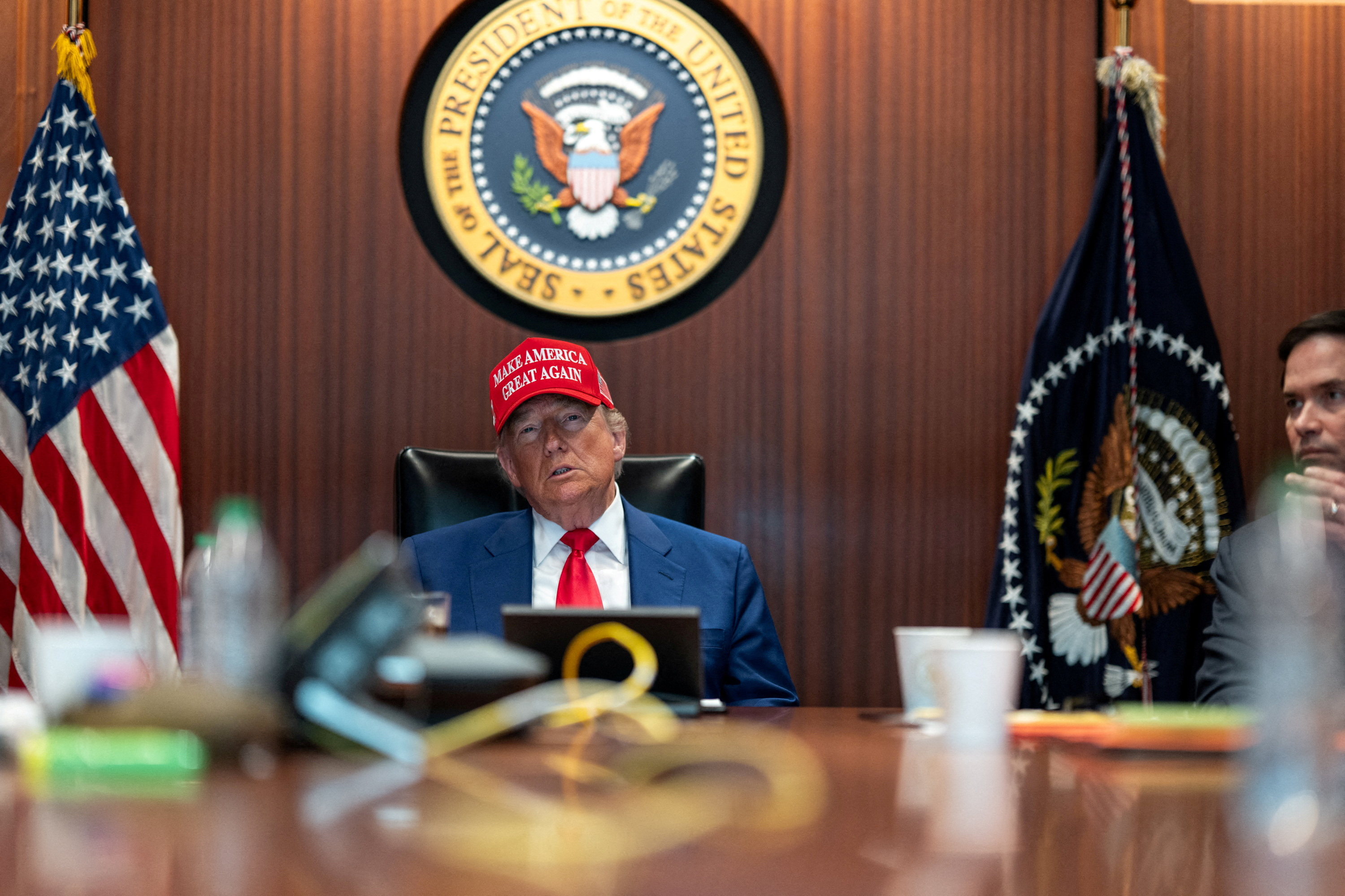 Donald Trump sits at a desk wearing a red Make America Great Again hat with a tablet in front of him, Secretary of State Marco Rubio next to him, and the presidential seal behind him.