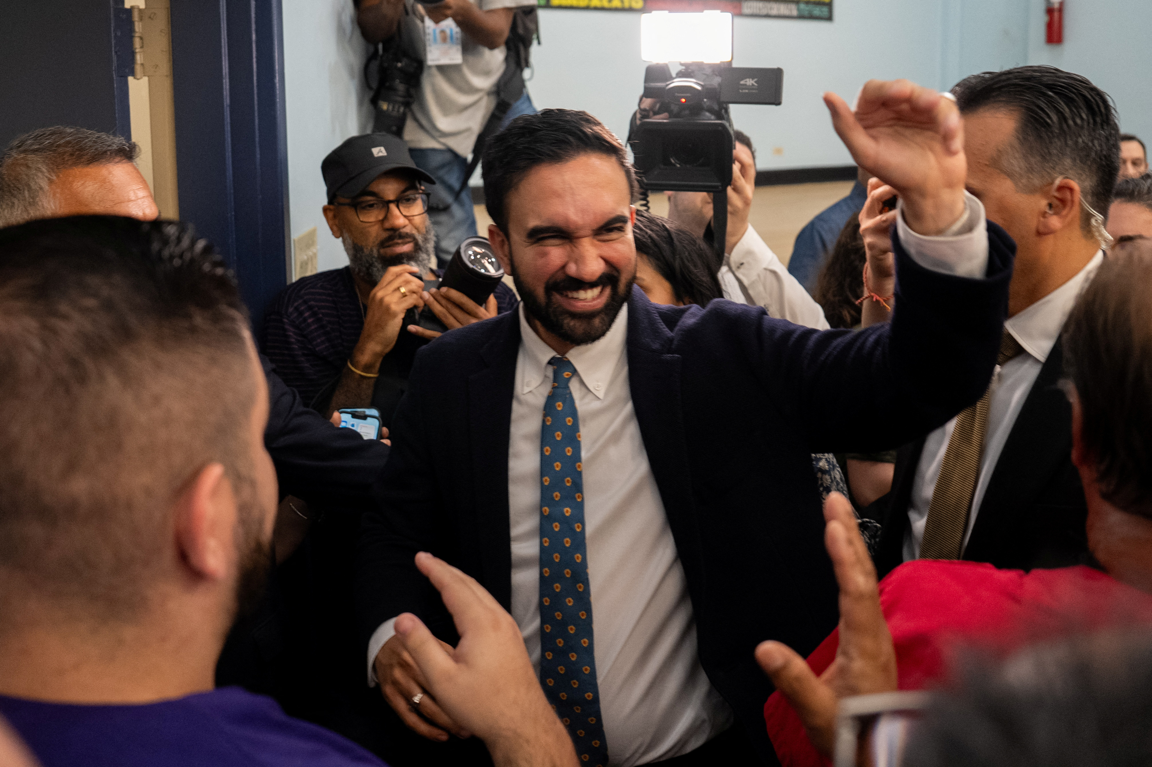 Zoran Mamdani waves as he leaves an event, surrounded by media cameras.
