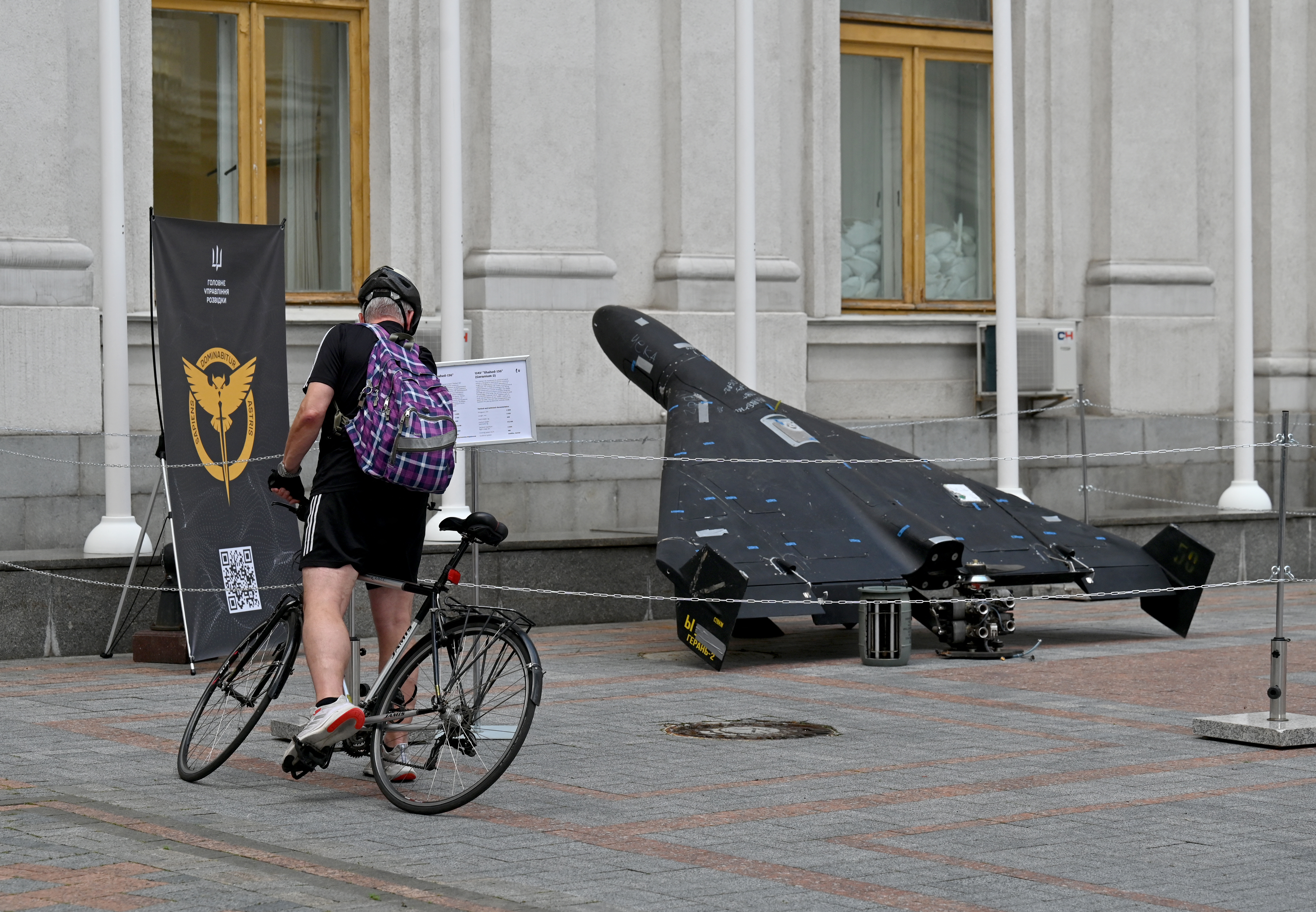 a man on a bicycle looks at a black drone outside an old building