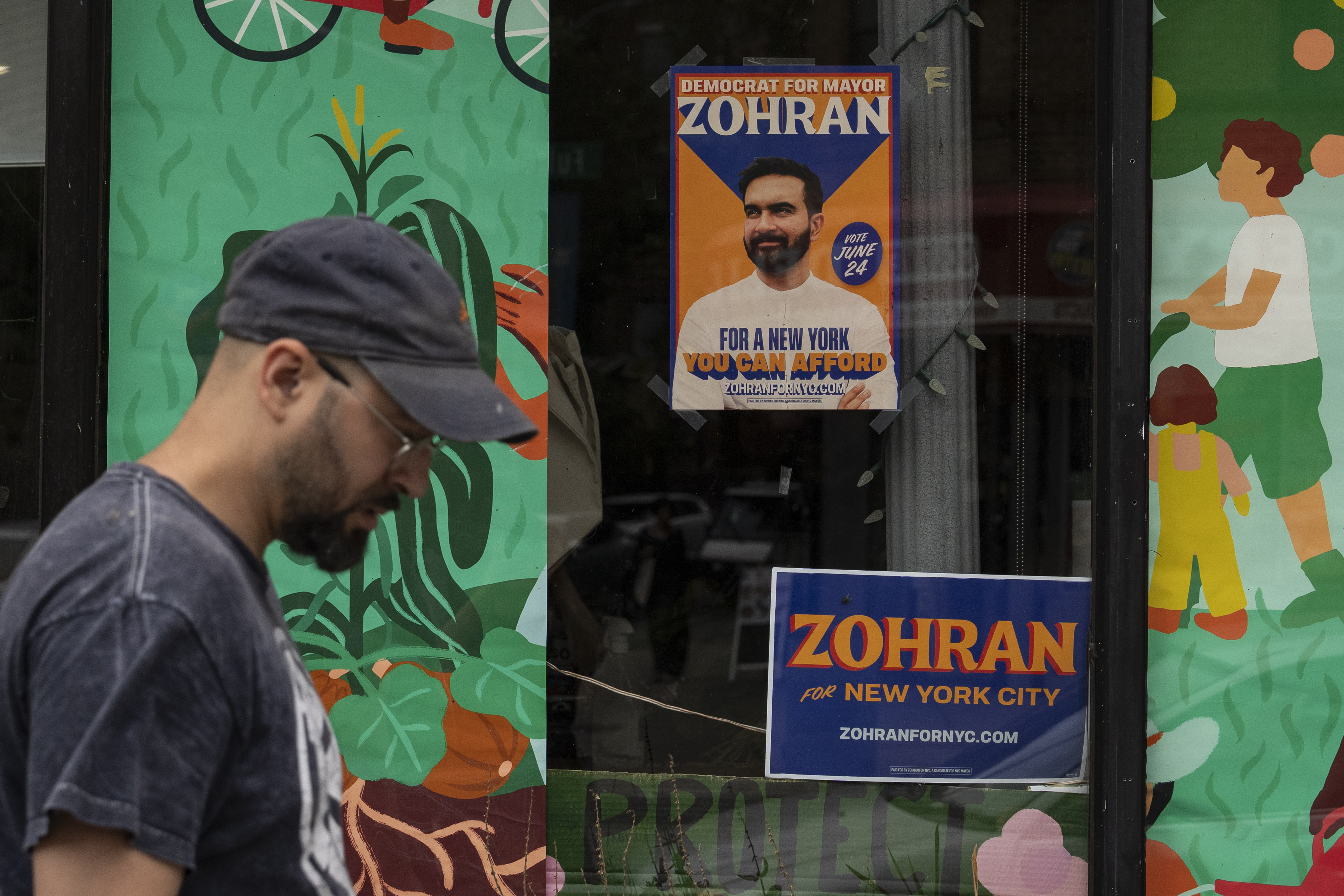 A pedestrian walks past two signs for Zohran Mamdani's mayoral campaign.
