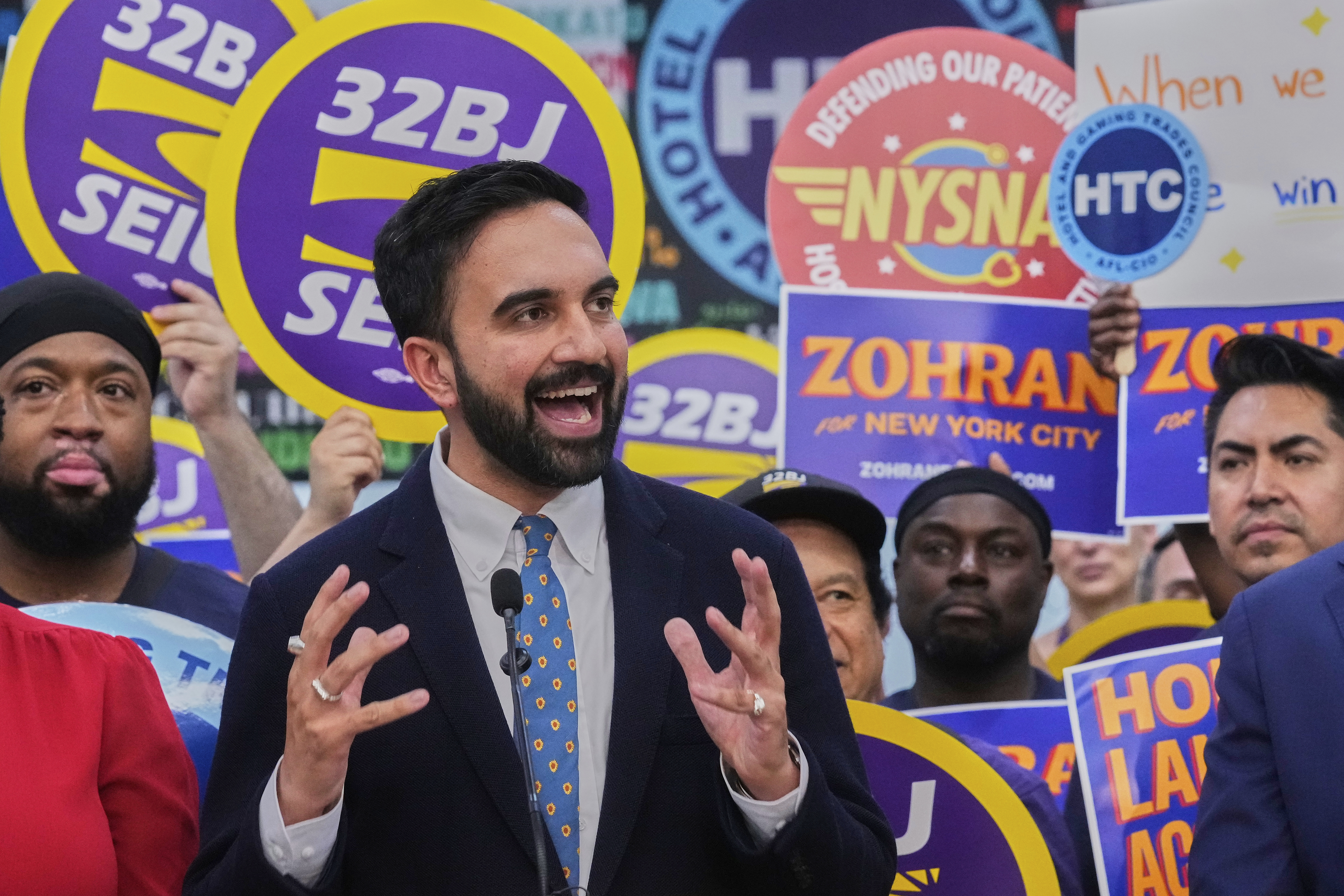 Zohran Mamdani at a rally, surrounded by supporters waving signs
