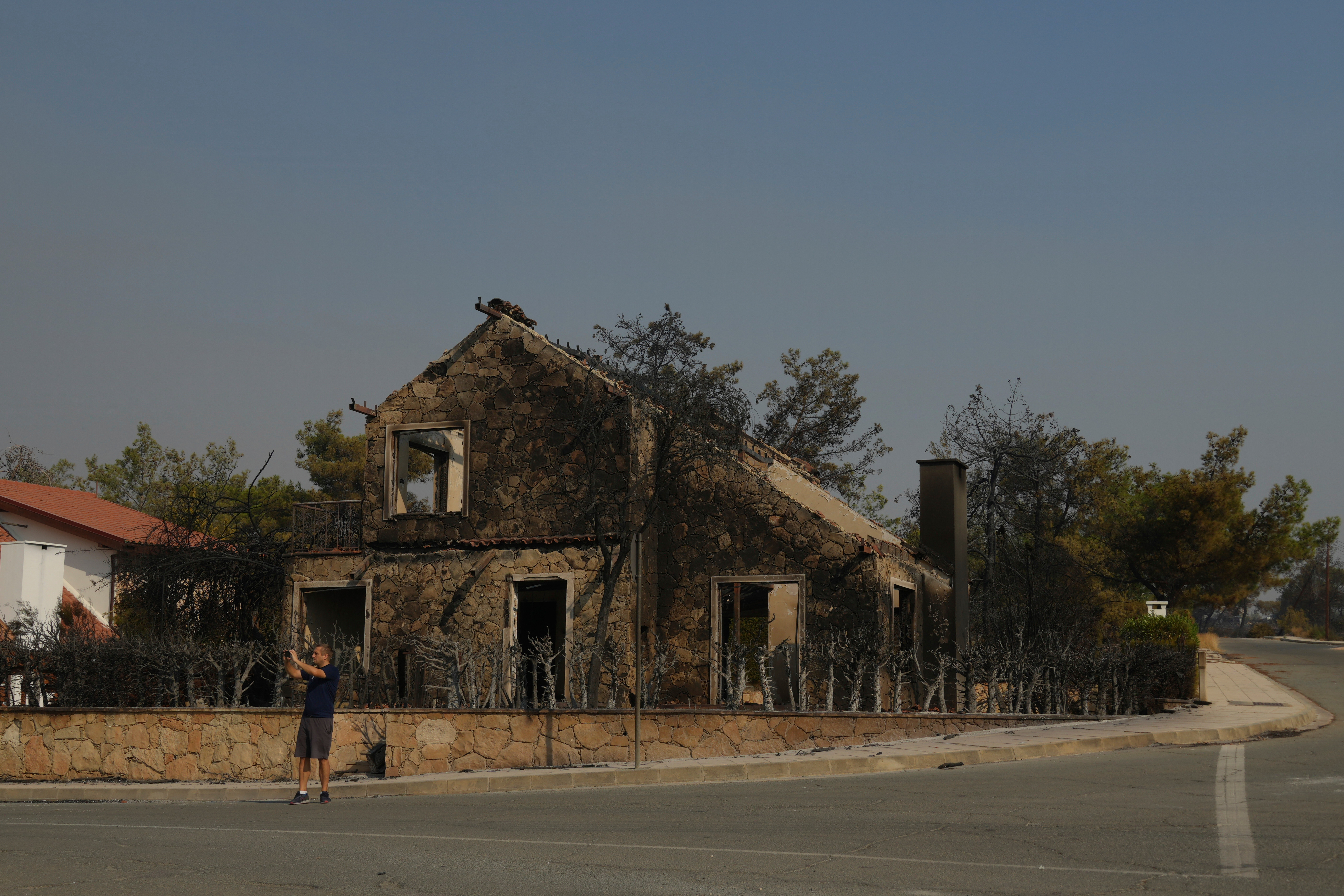 A man takes a photograph in front of a burned house in Souni village, Cyprus, during a massive wildfire on the southern side of the east Mediterranean island nation's Troodos mountain range, Thursday, July 24, 2025. (AP Photo/Petros Karadjias)