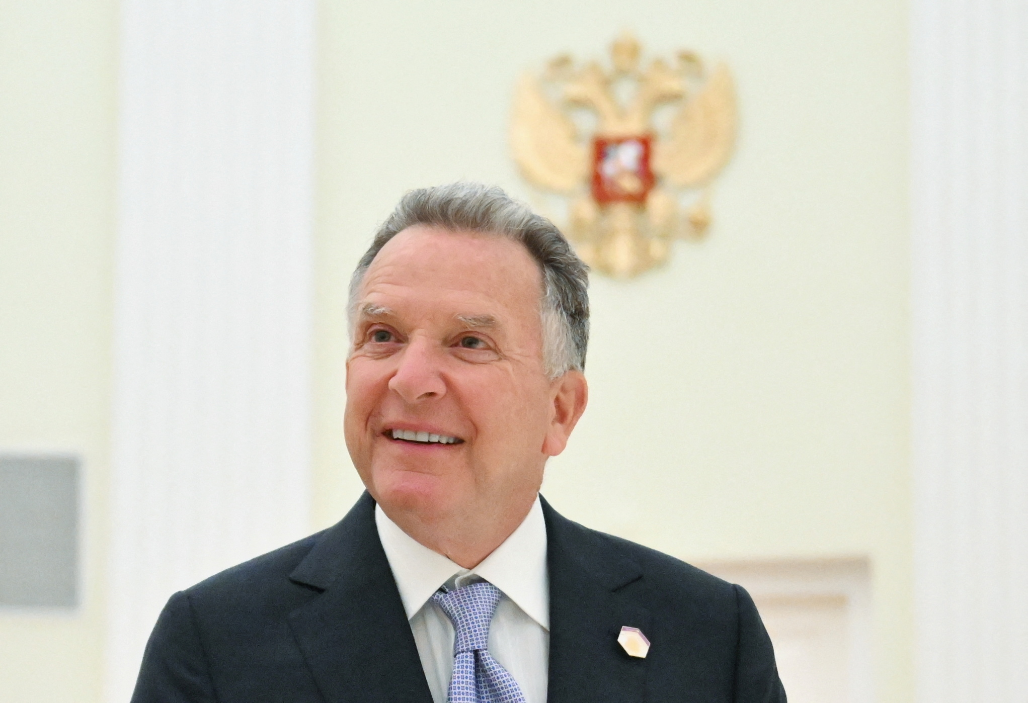 a man in a suit in front of a gold eagle crest on a white wall