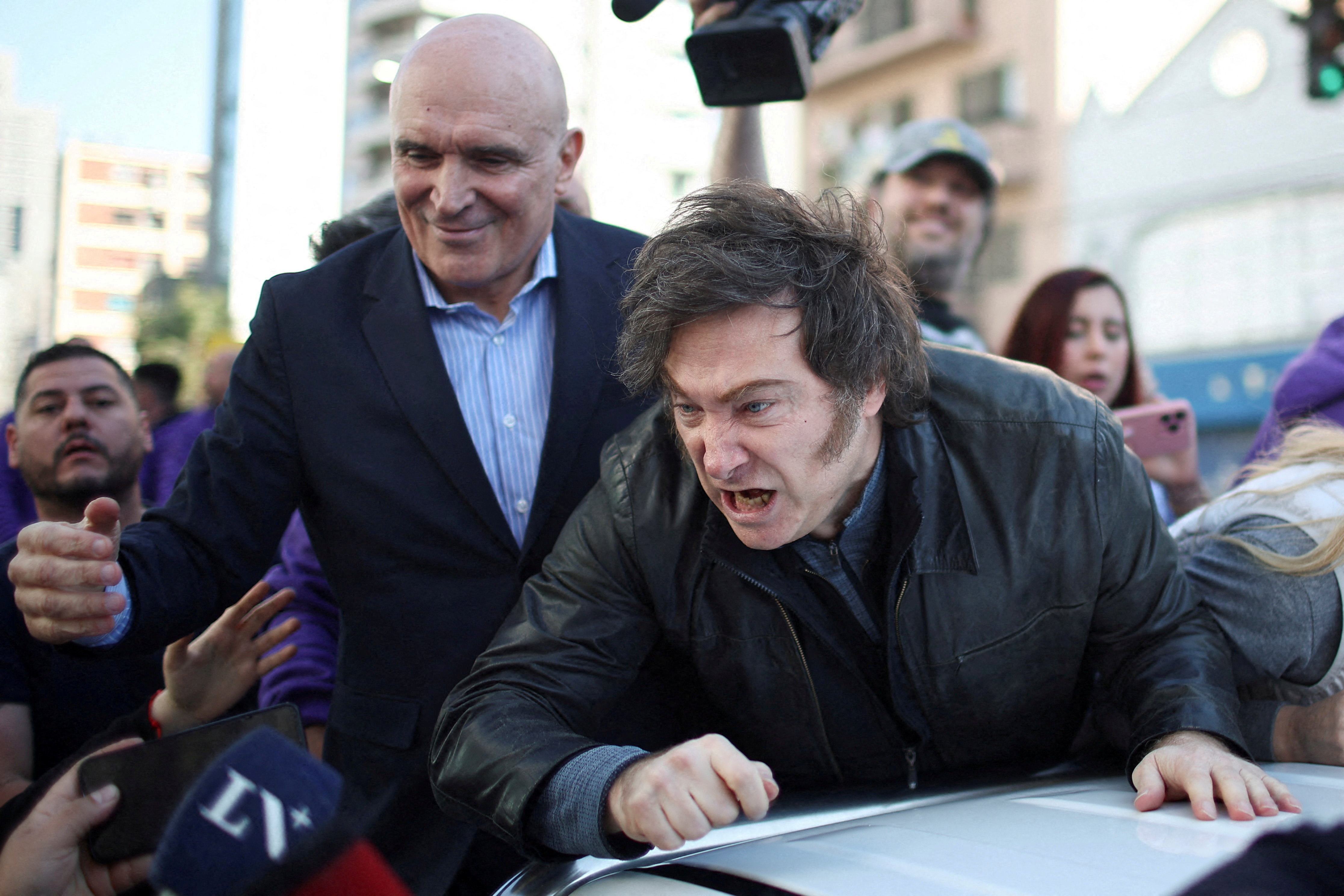 Javier Milei leans over a car to speak to a reporter's microphone.