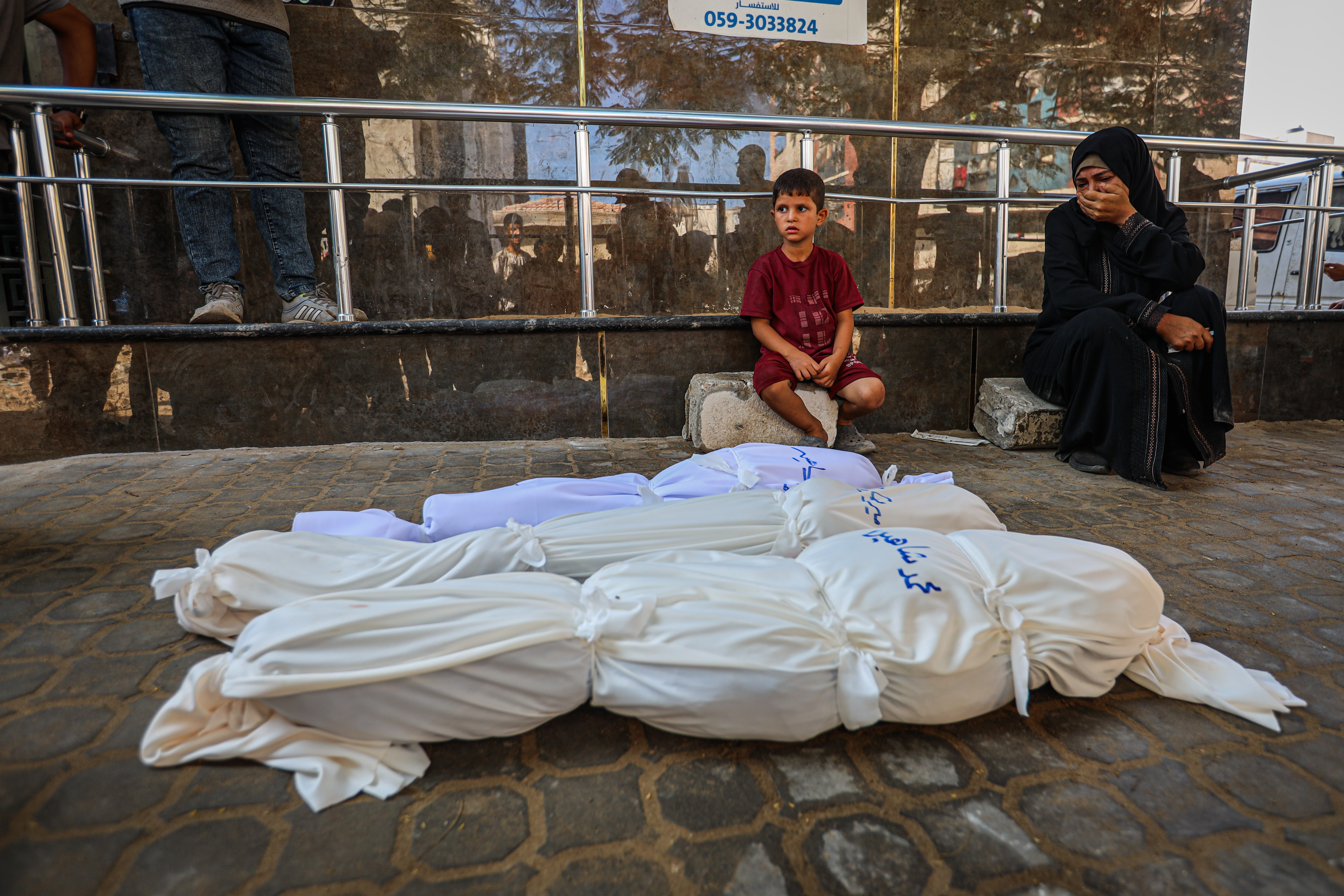 GAZA CITY, GAZA - AUGUST 22: A grieving woman sits next to a young boy outside Al-Shifa Hospital as white-shrouded bodies of Palestinians killed in an Israeli airstrike lie on the ground awaiting funeral arrangements in Gaza City on August 22, 2025. Photojournalist:Hamza Z. H. Qraiqea