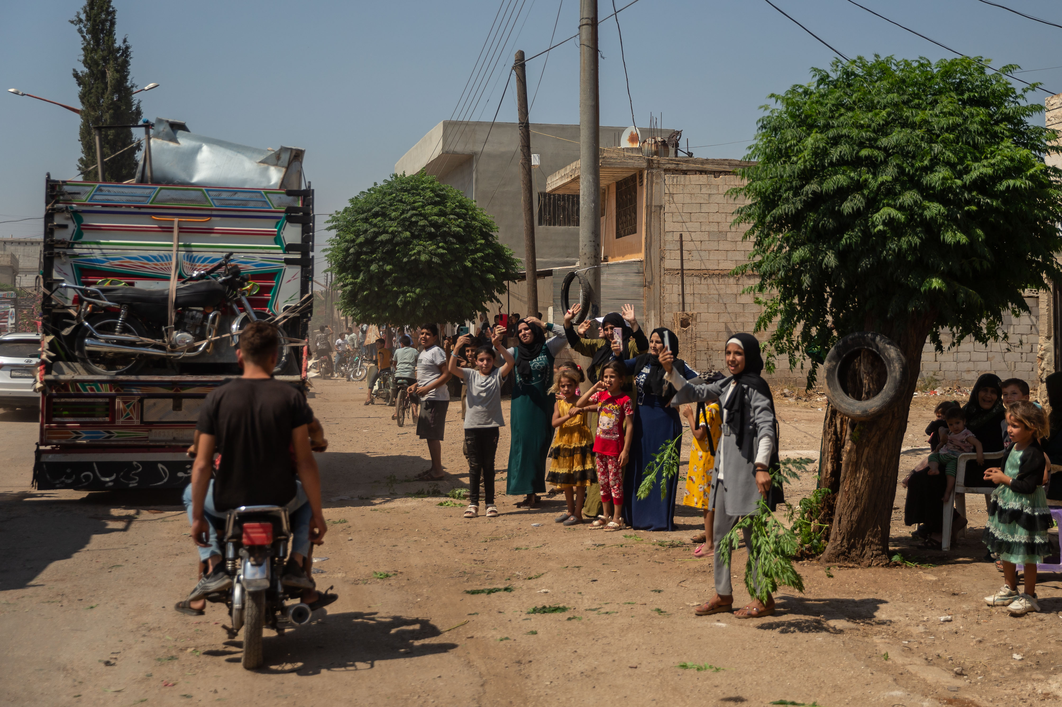 A line of people waiting on the side of a dusty road wave as a truck carrying returnees, as well as a motorbike, pass by