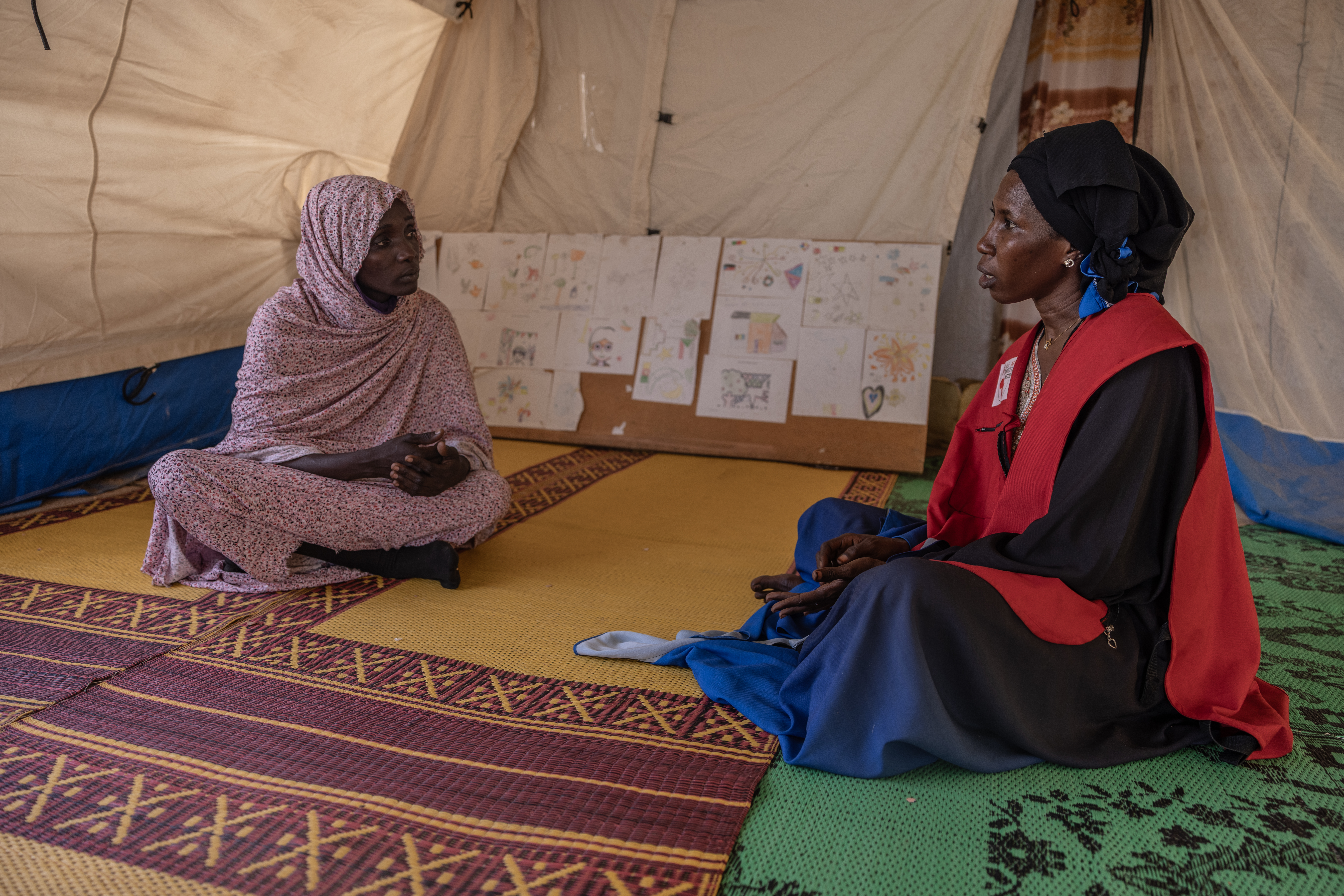 Two women sit on plastic mats on the floor of a tent, counselor on the right and a refugee