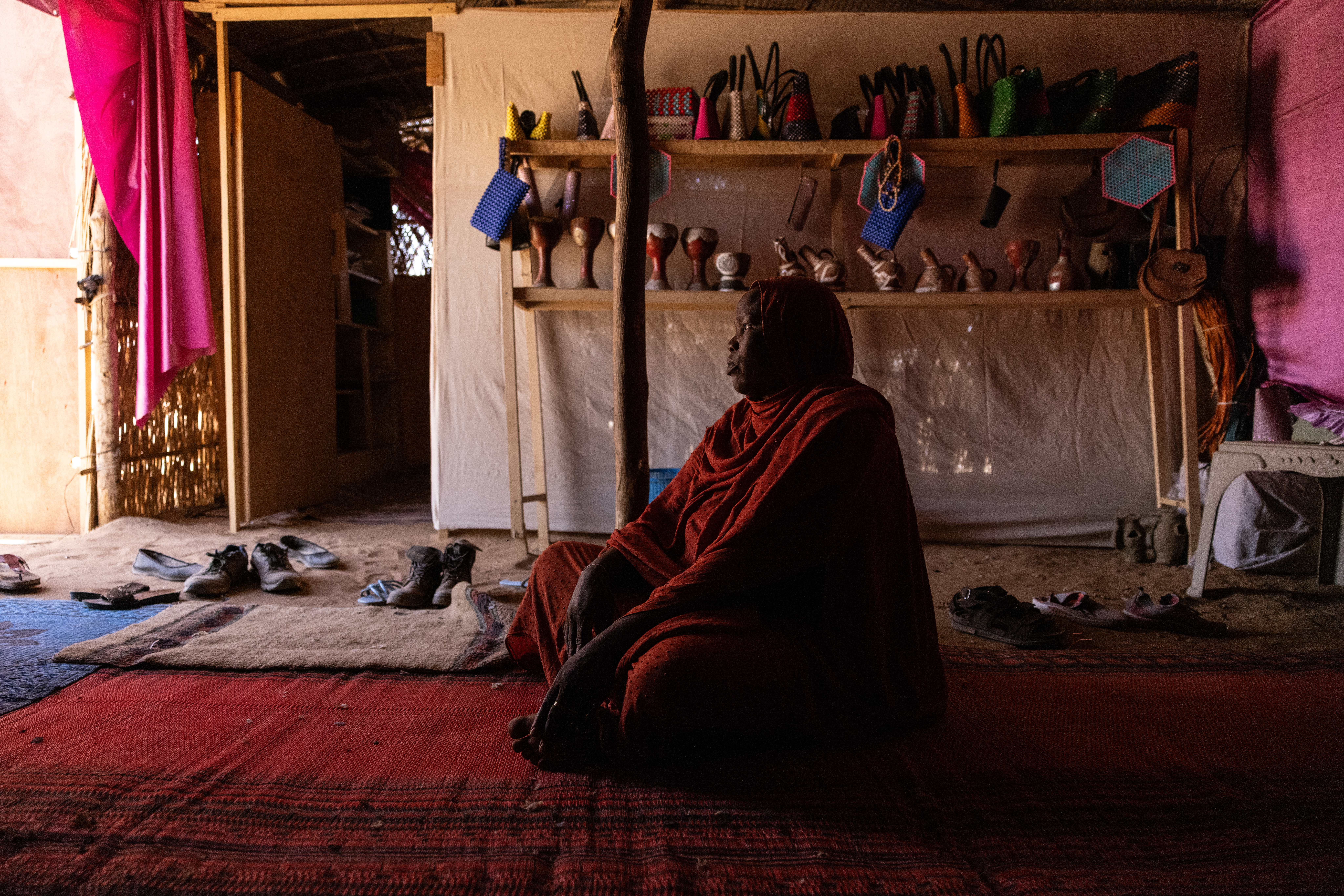 A woman in a dark red thobe sits on the ground in a dimply lit tent