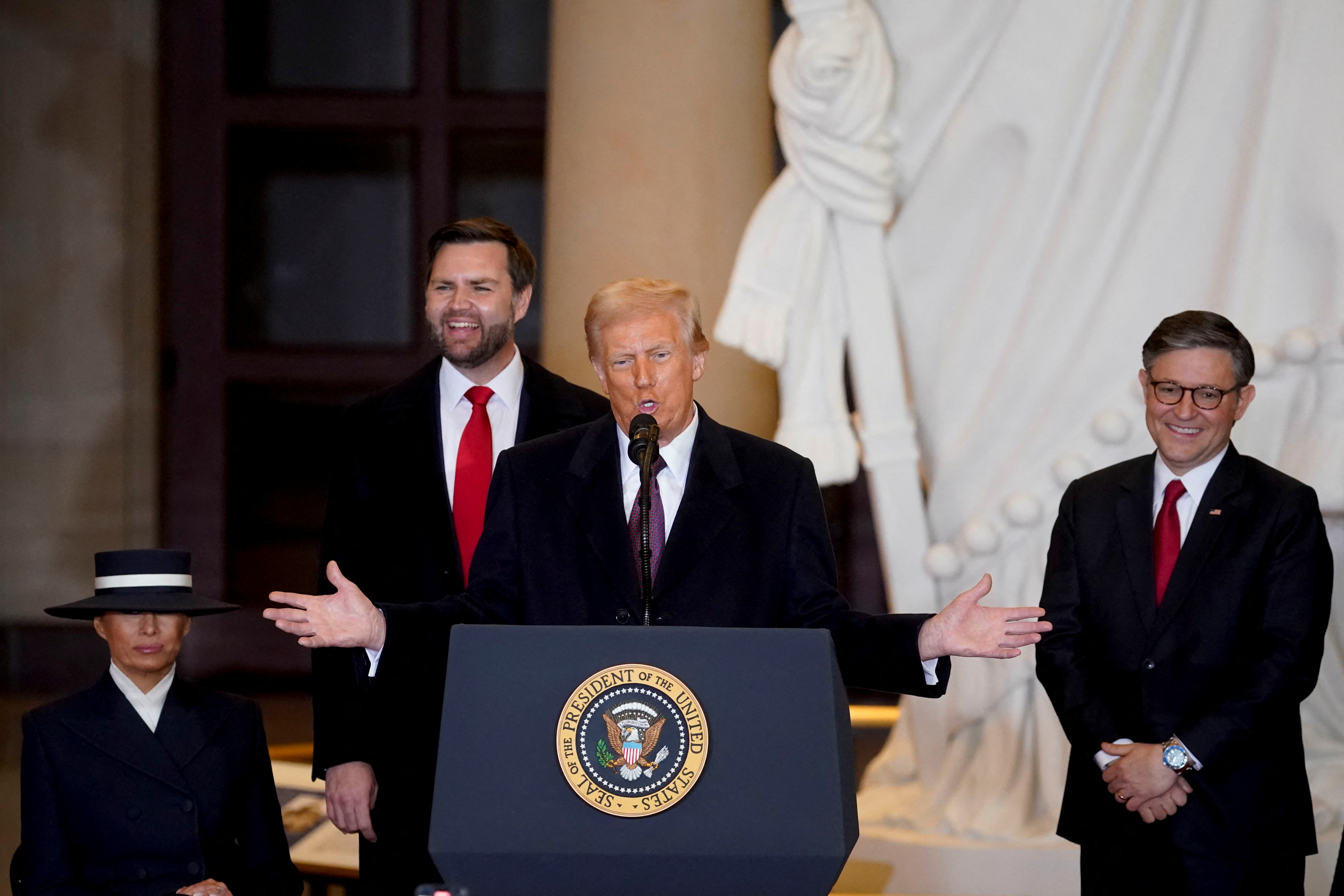 Donald Trump gives an inauguration speech behind a presidential podium, next to Mike Johnson, JD Vance and Melania Trump. A marble statue is visible behind them.