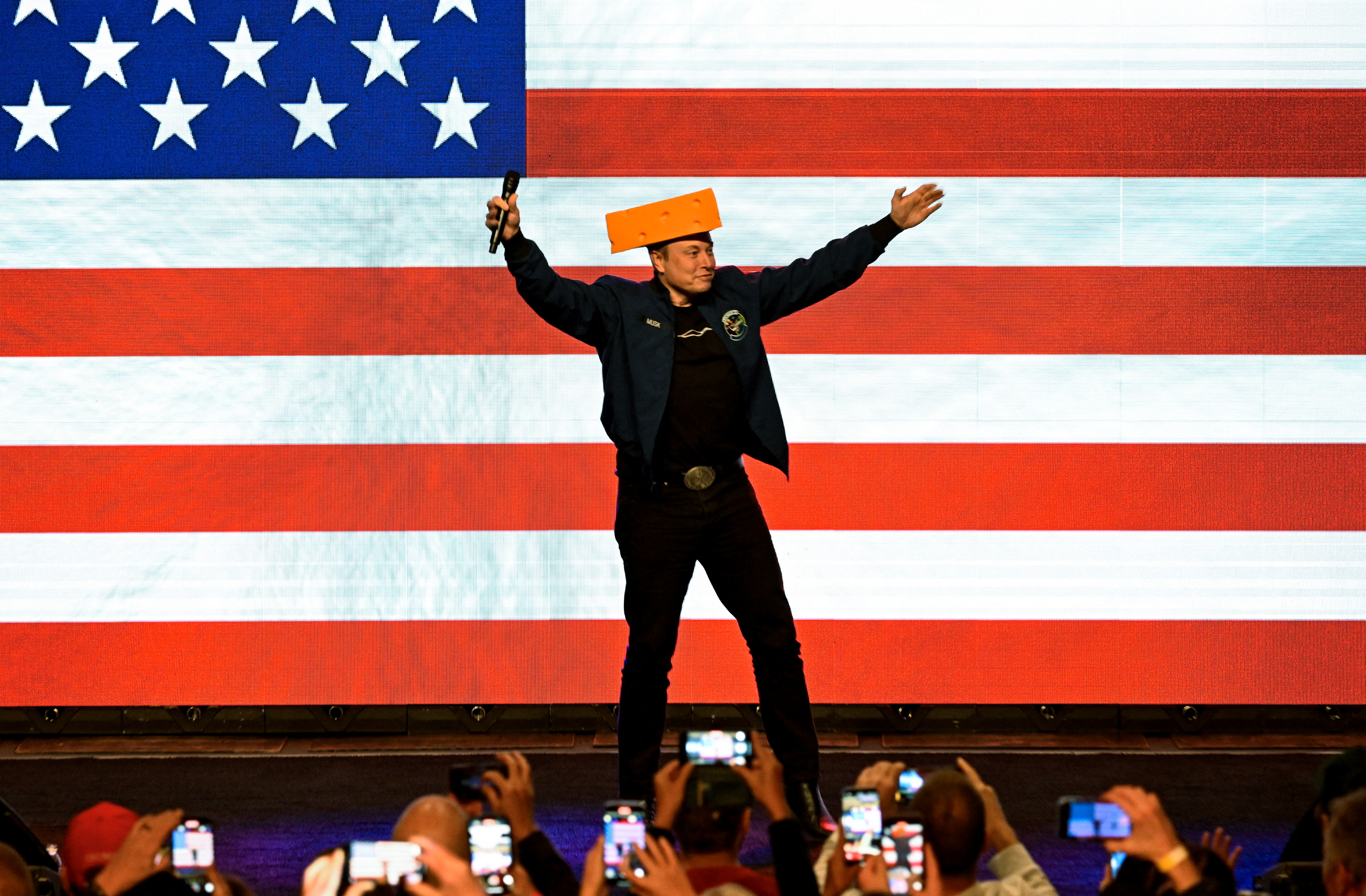 Elon Musk in a cheese hat stands on stage against a US flag