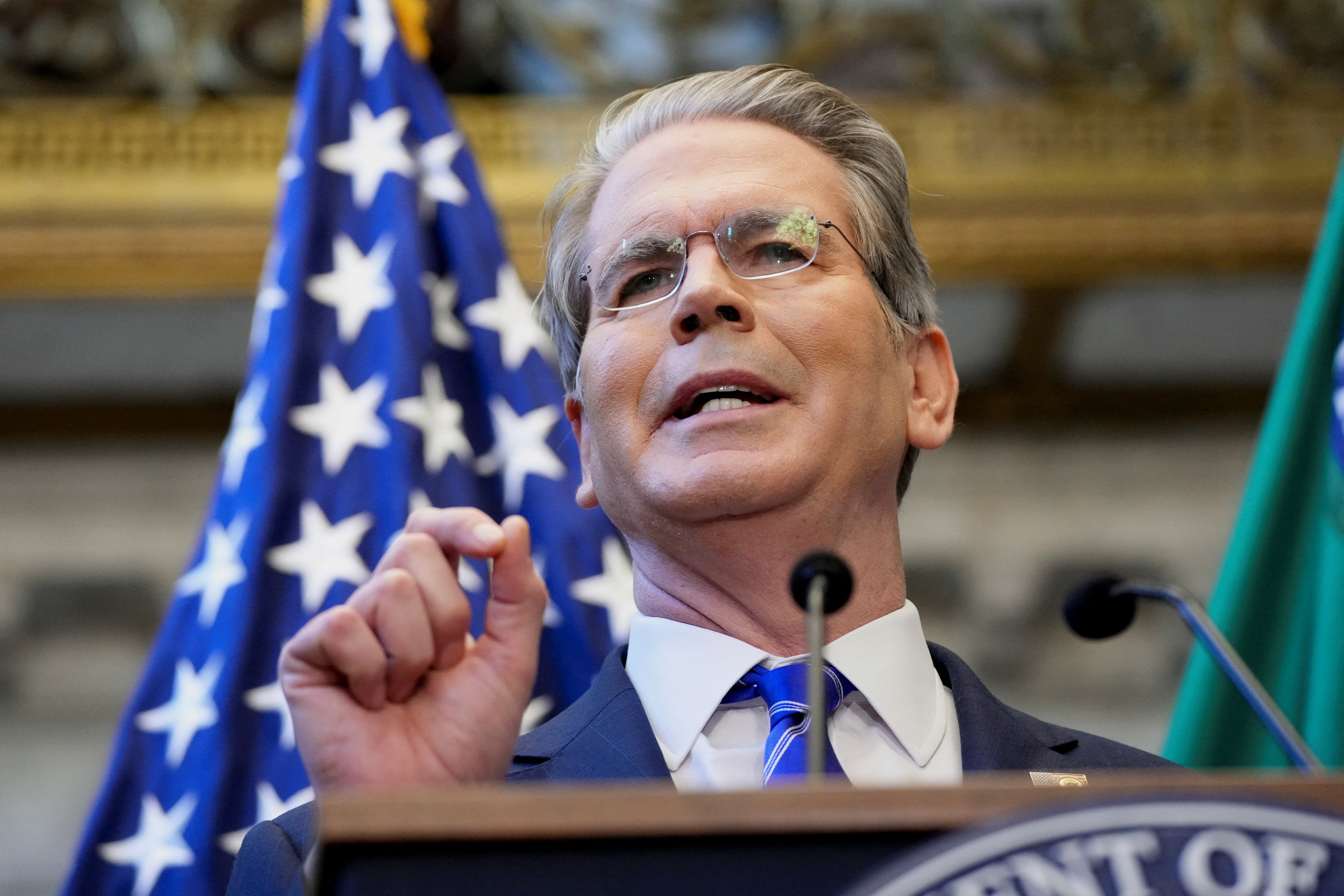 Treasury Secretary Scott Bessent stands behind a podium speaking, in front of a US flag.
