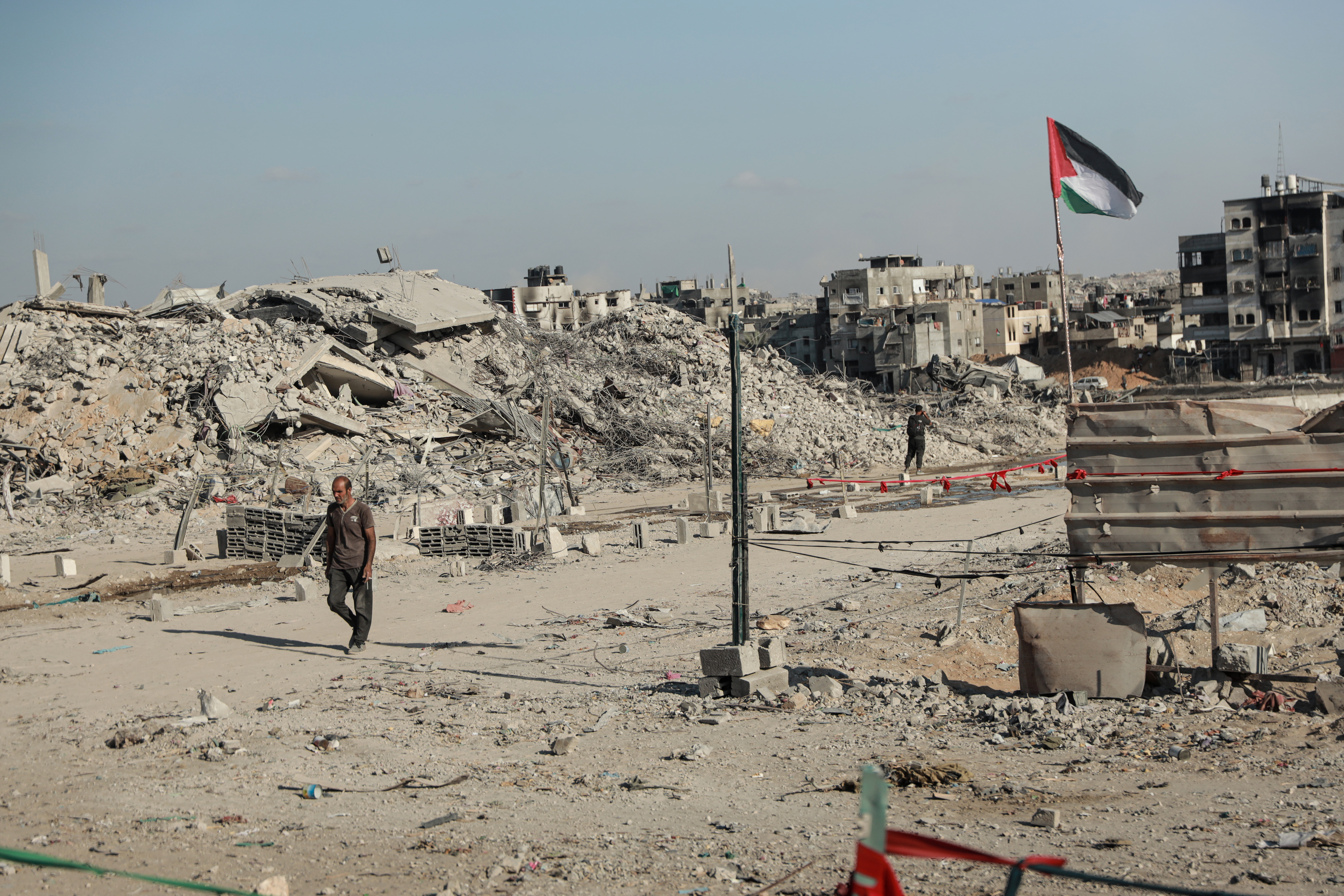 A man walks with rubble and a Palestinian flag in the background