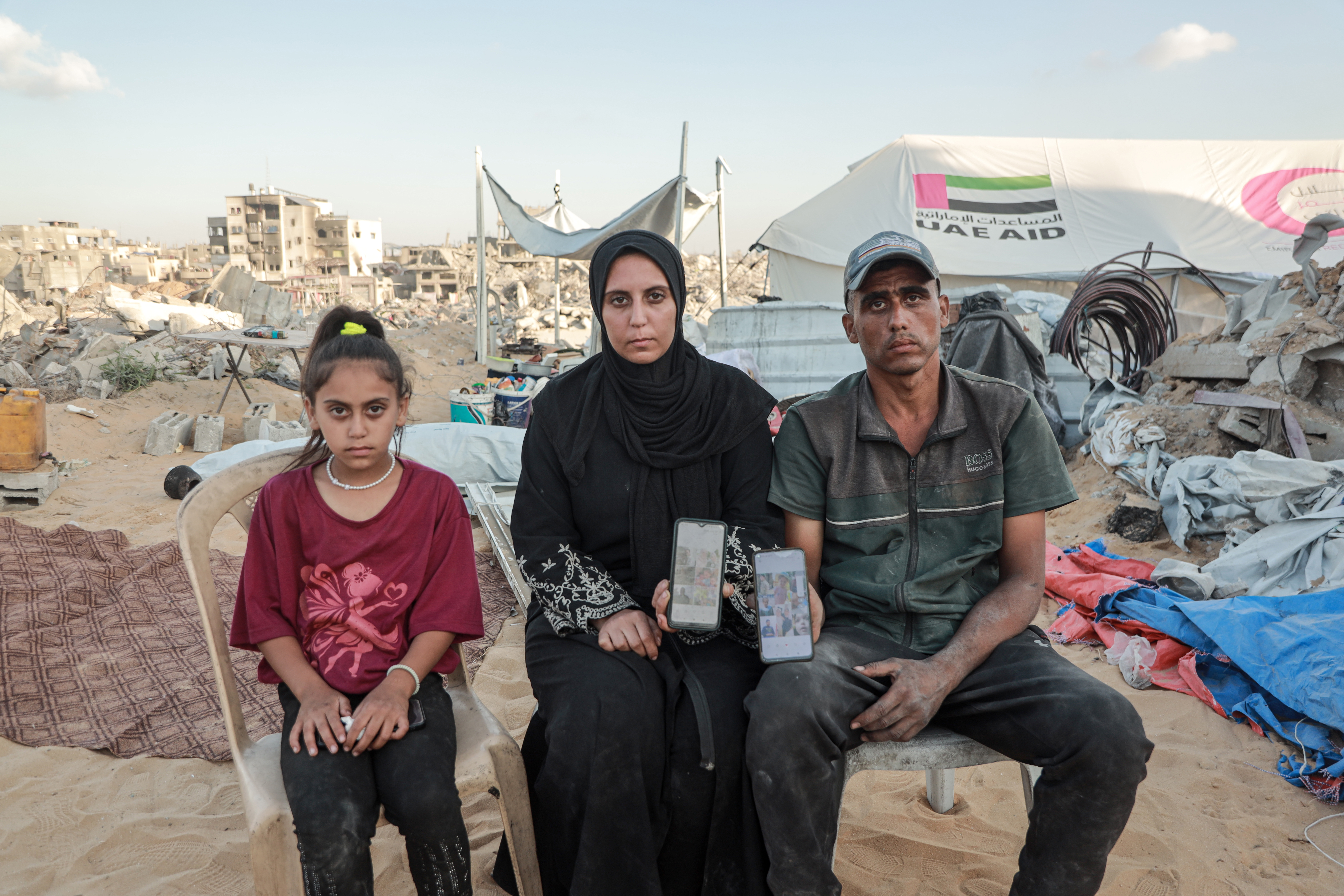 A girl, her mother, and her father sit on chairs next to tents and rubble