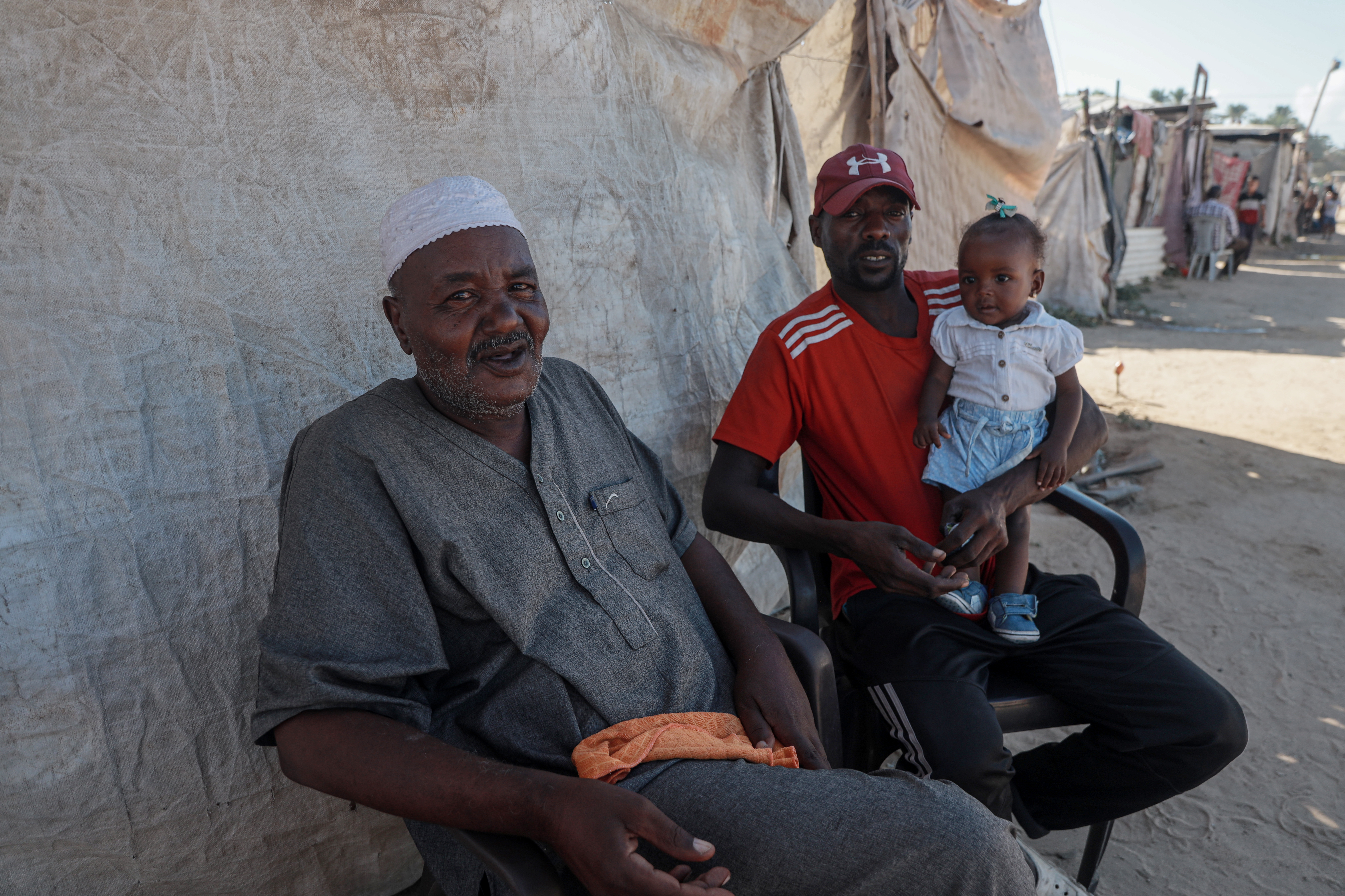 An elderly man smiles at the camera, he's sitting on a plastic chair by an old wall. he looks tired and emaciated
