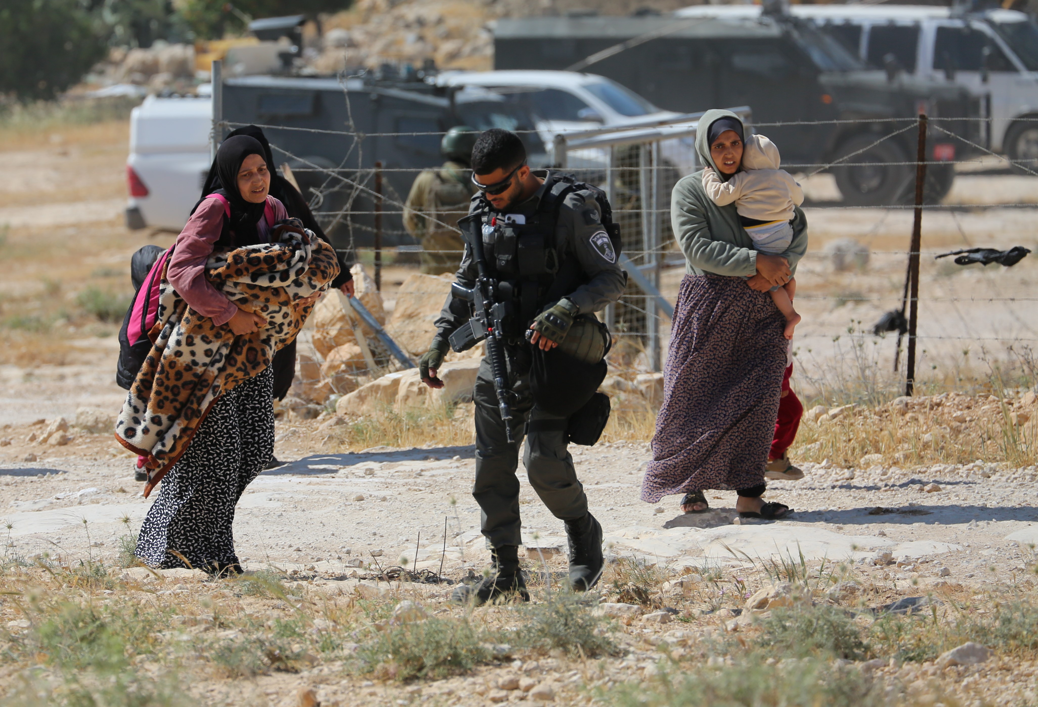 Two Palestinian women carrying children as they walk behind an Israeli soldier