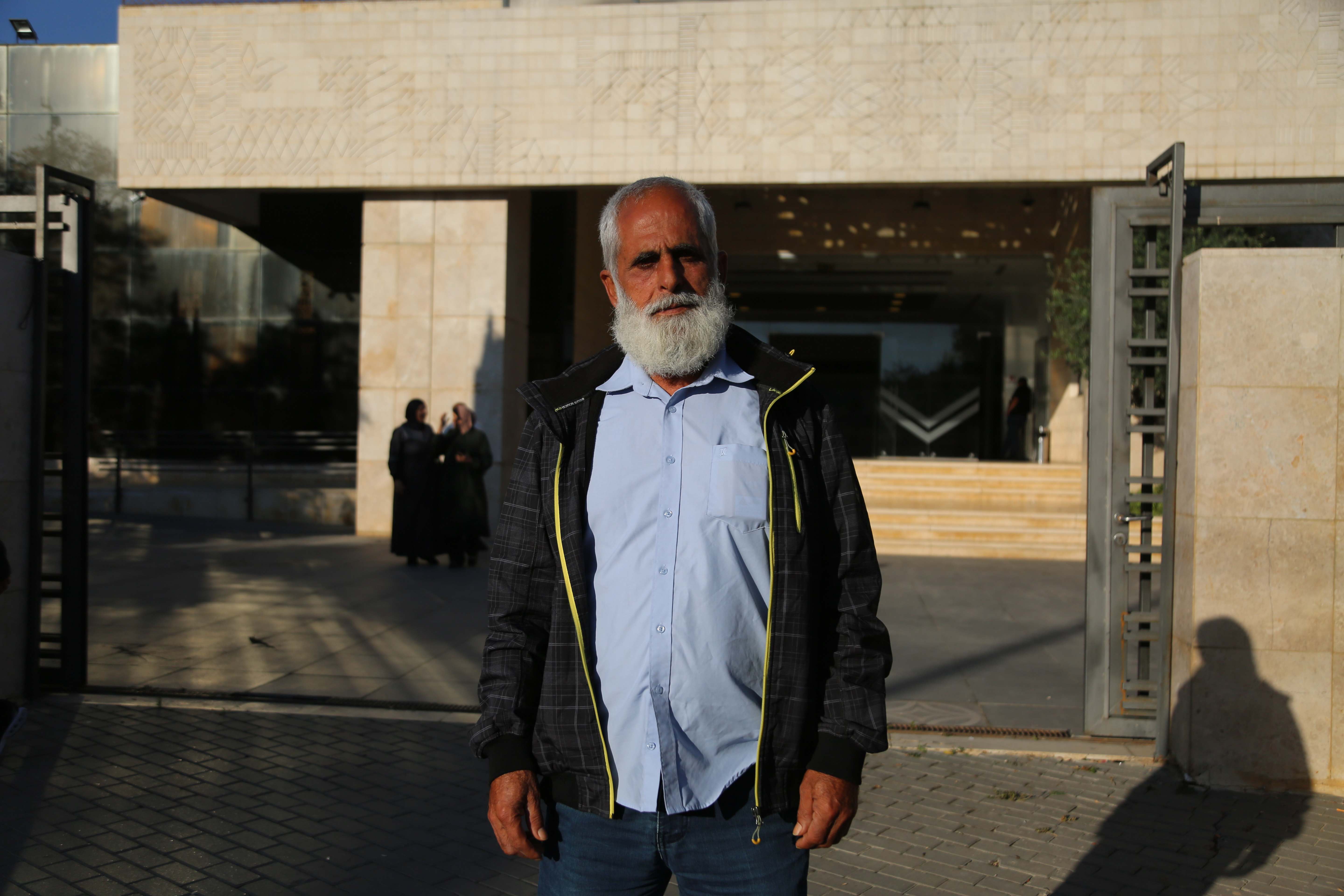 A kindly looking man with a soft grey beard, wearing a tidy windbreaker and oxford cloth shirt looks traight at the camera