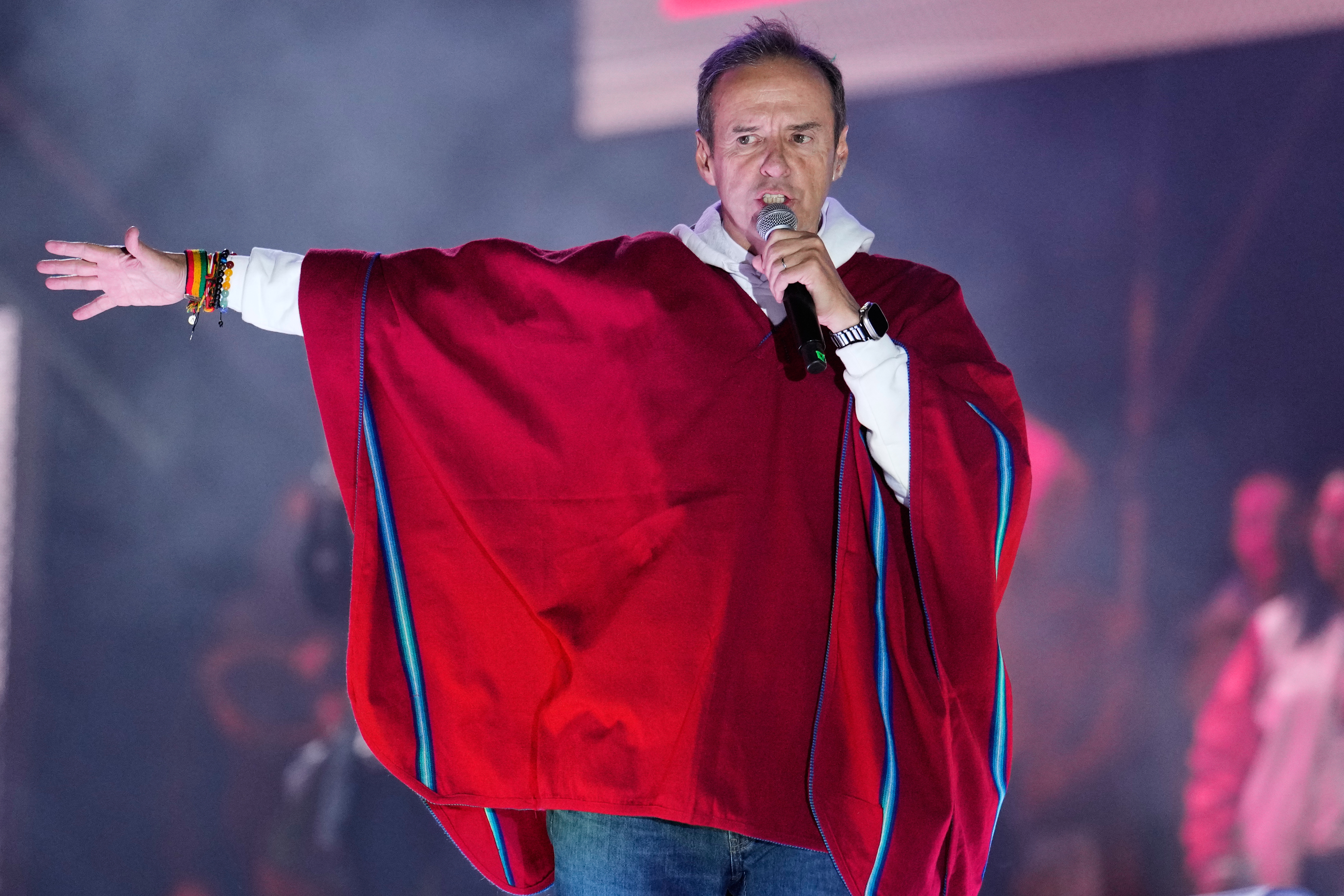 FOR BRIAN OSGOOD Jorge Quiroga wears a red poncho and speaks into a microphone on stage, one arm outstretched.
