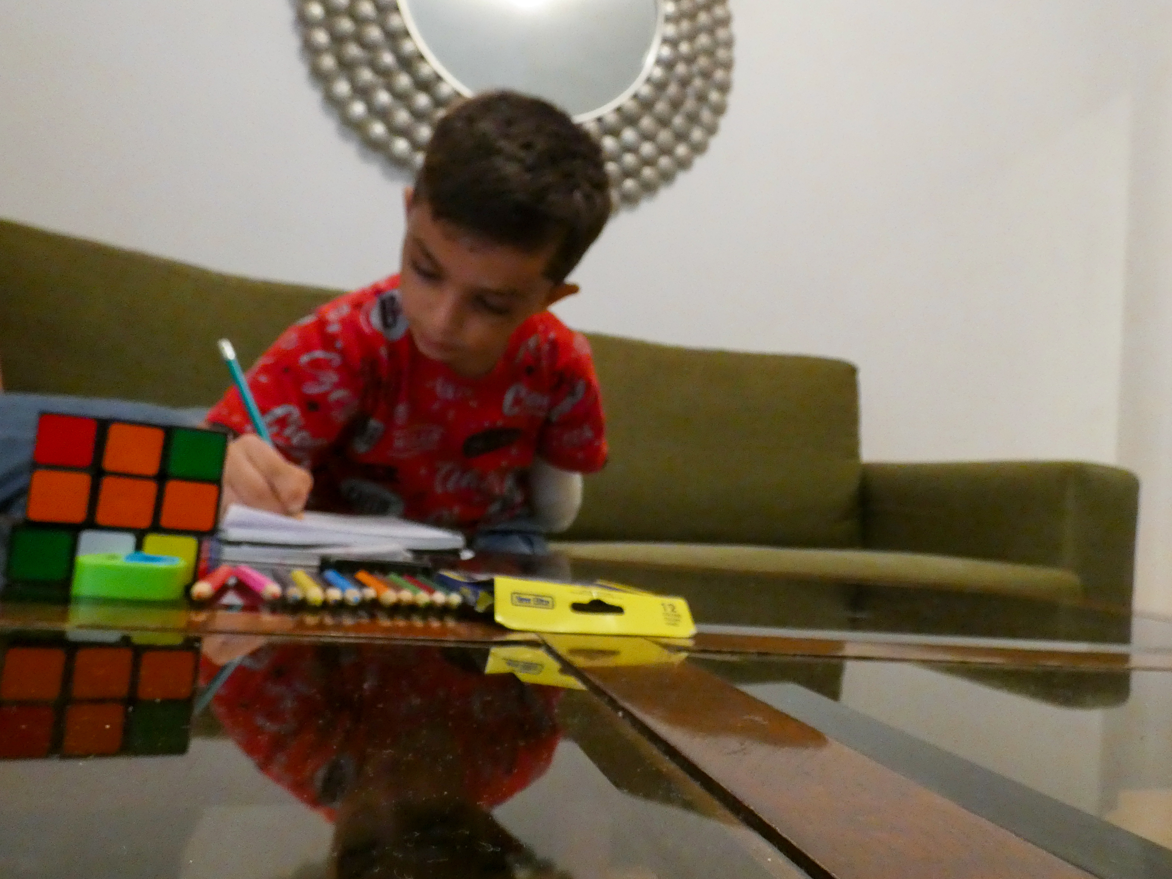 A little boy in a red t-shirt leans over, drawing with his right hand. His left arm is a stump, tucked by his side. In front of him are some coloured pencils and a Rubiks Cube