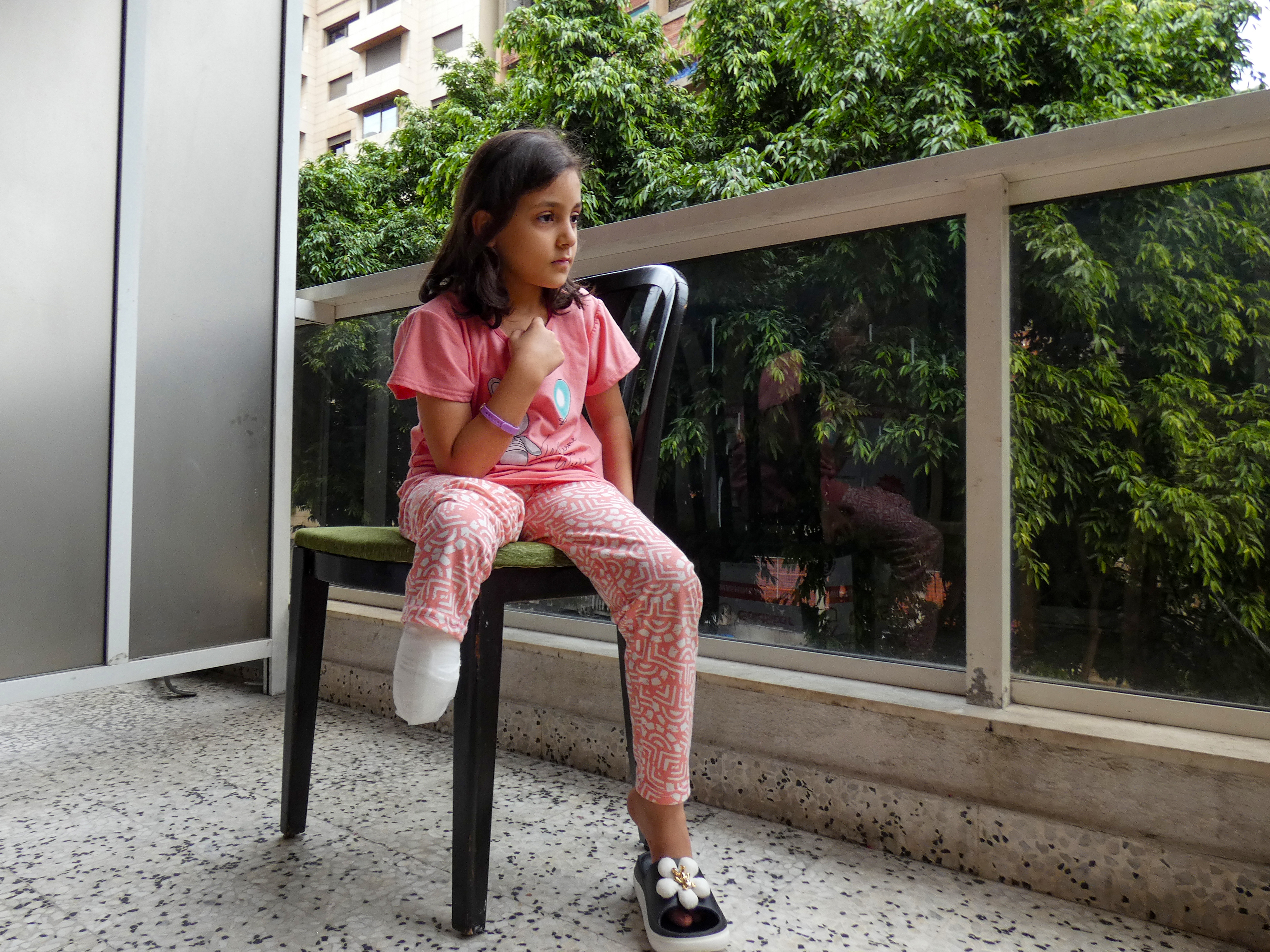 A little girl in pink pajamas sits on a plastic chair in a balcony, one plastic slipper with a flower on her left foot. Her right leg is amputated, a stump hangs below her knee.